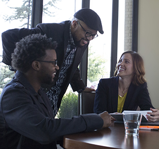 Two people sit in an office talking to a man standing behind them.