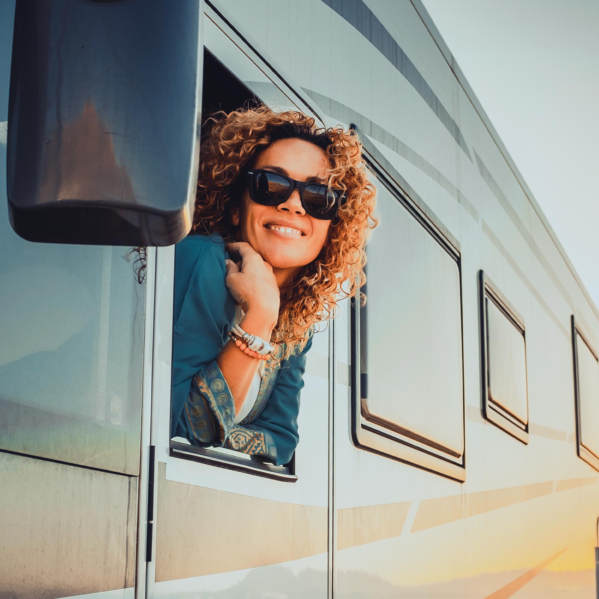 A woman with sunglasses hangs out the window of her motorhome smiling.