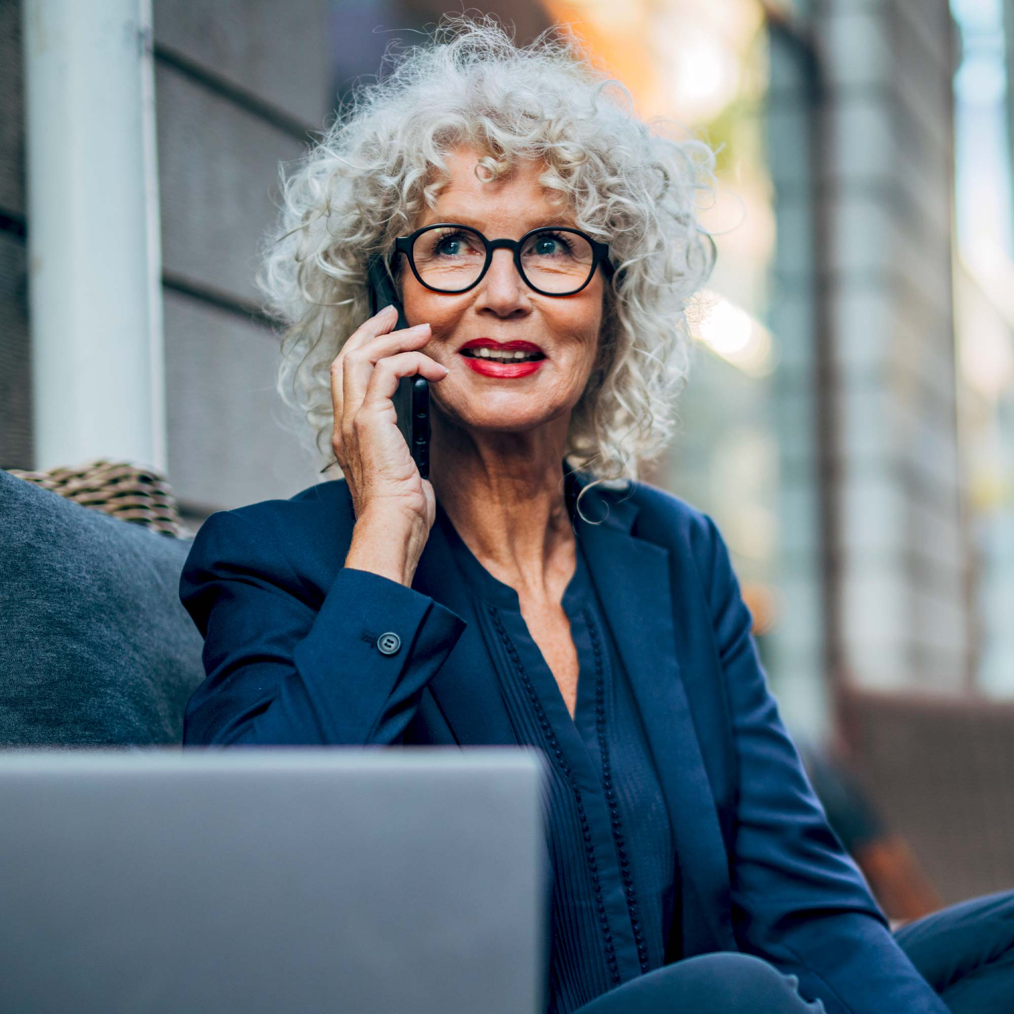 A woman in a blue business jacket talks on her cell phone in front of her laptop.