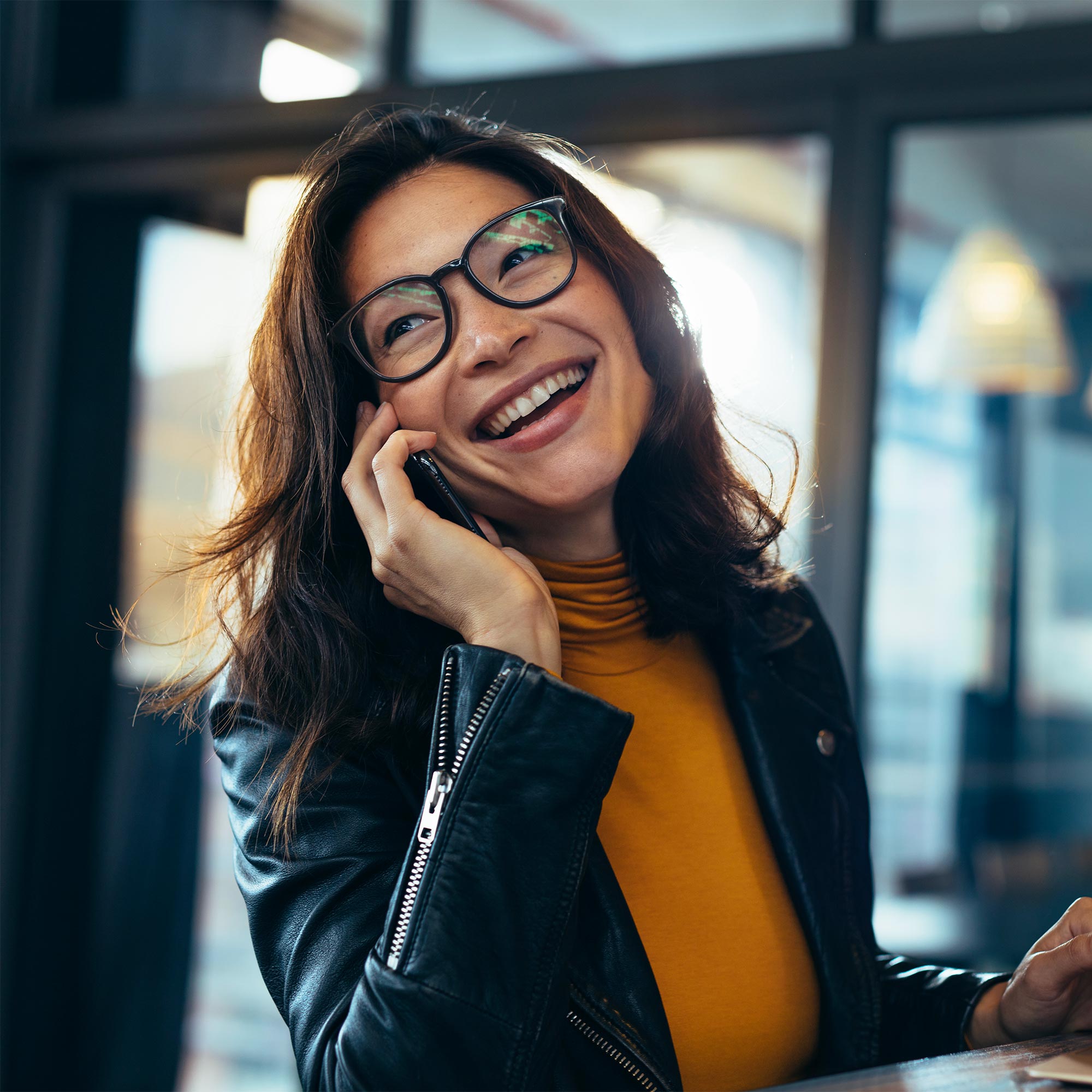 A woman wearing a black jacket and yellow shirt smiles as she talks on the phone.