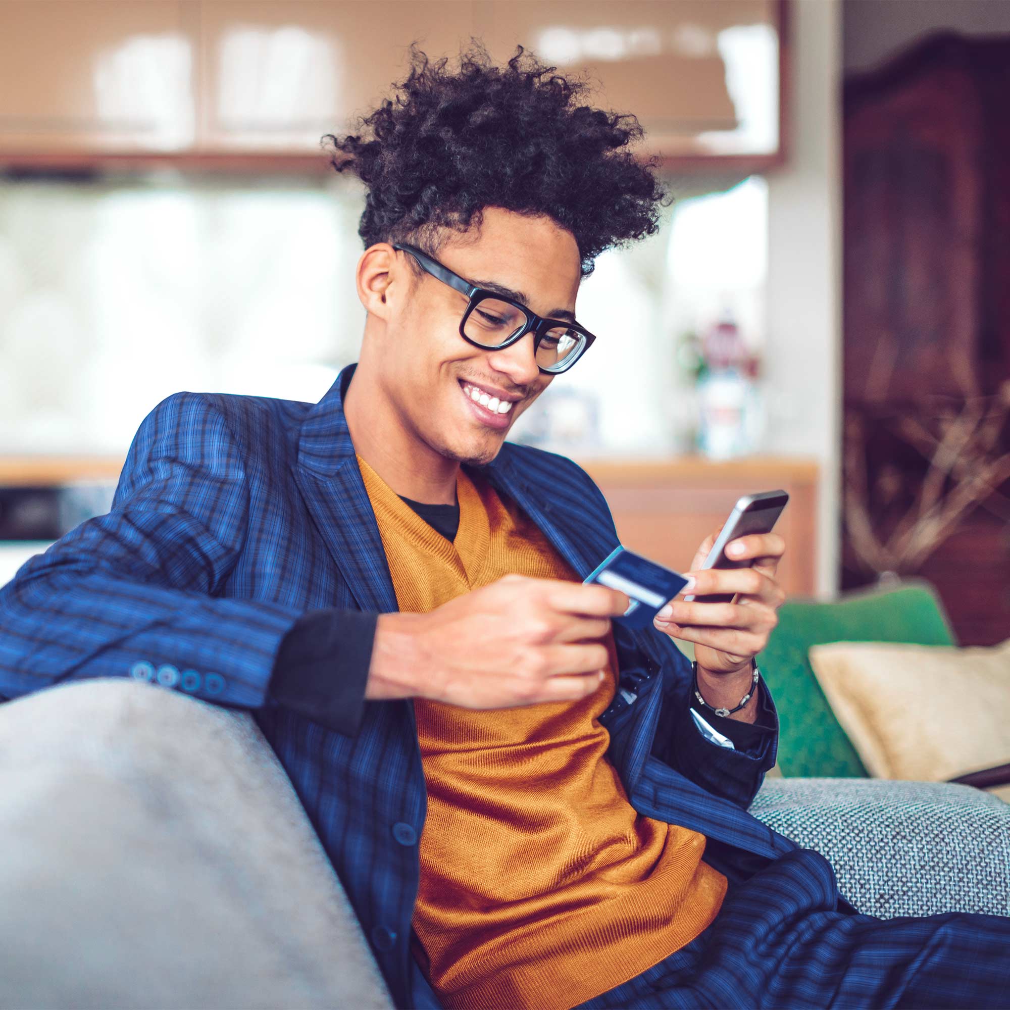 A man sits on the couch holding his debit card and phone and smiles.