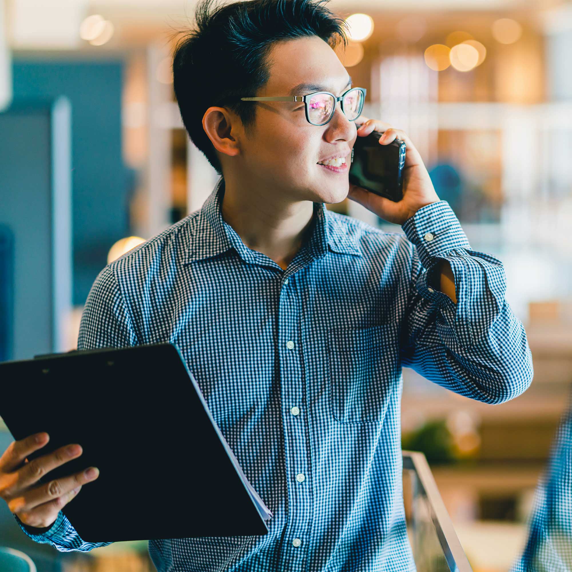 A man in a denim shirt holds his phone to his ear with one hand and has a notebook in the other.
