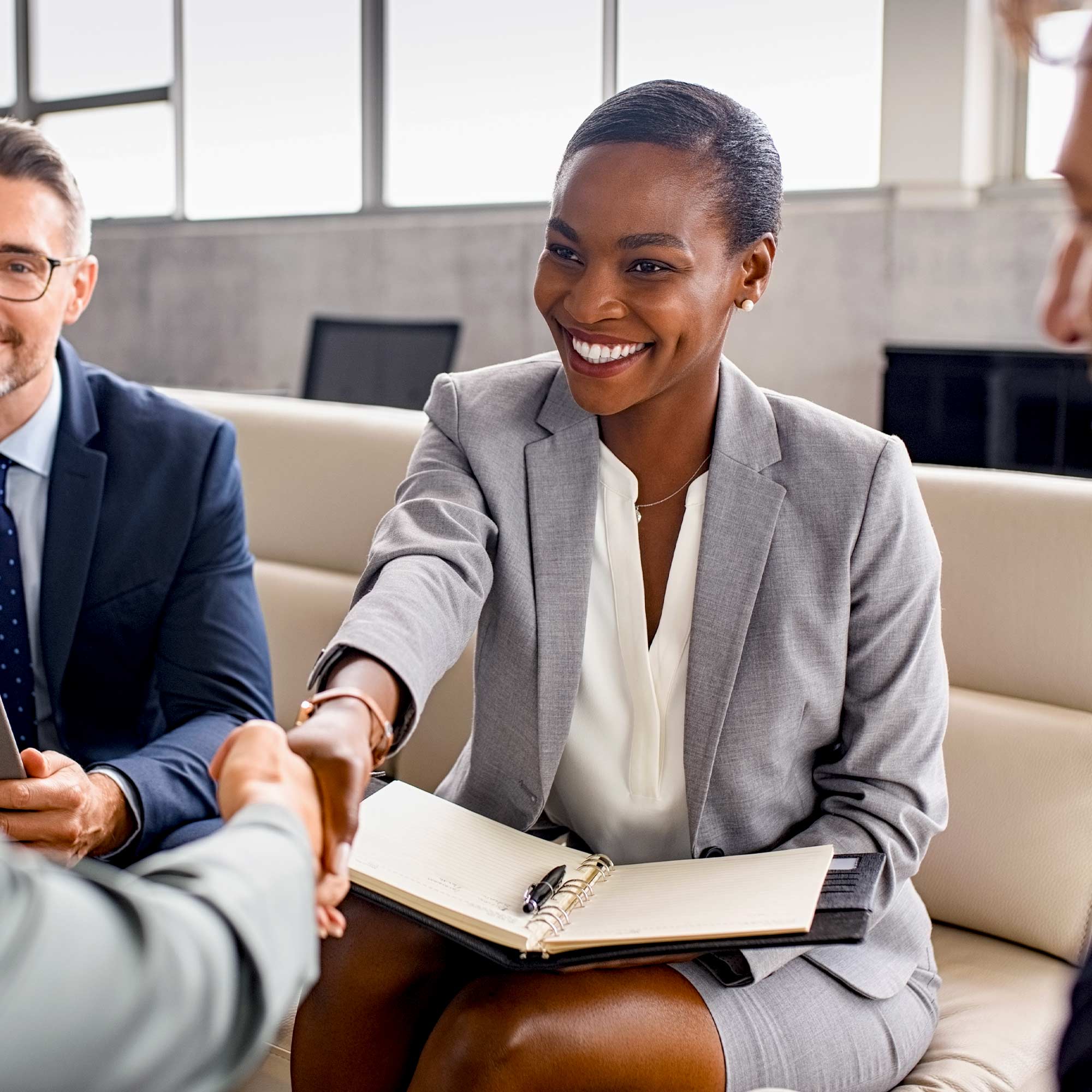 A professionally dressed woman sits on a couch and shakes hands with someone.