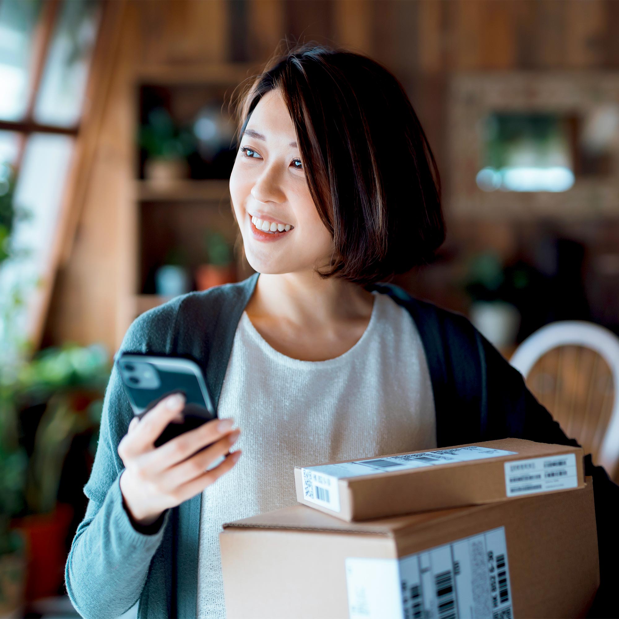 A woman stands behind a stack of packages with her phone in her hand.