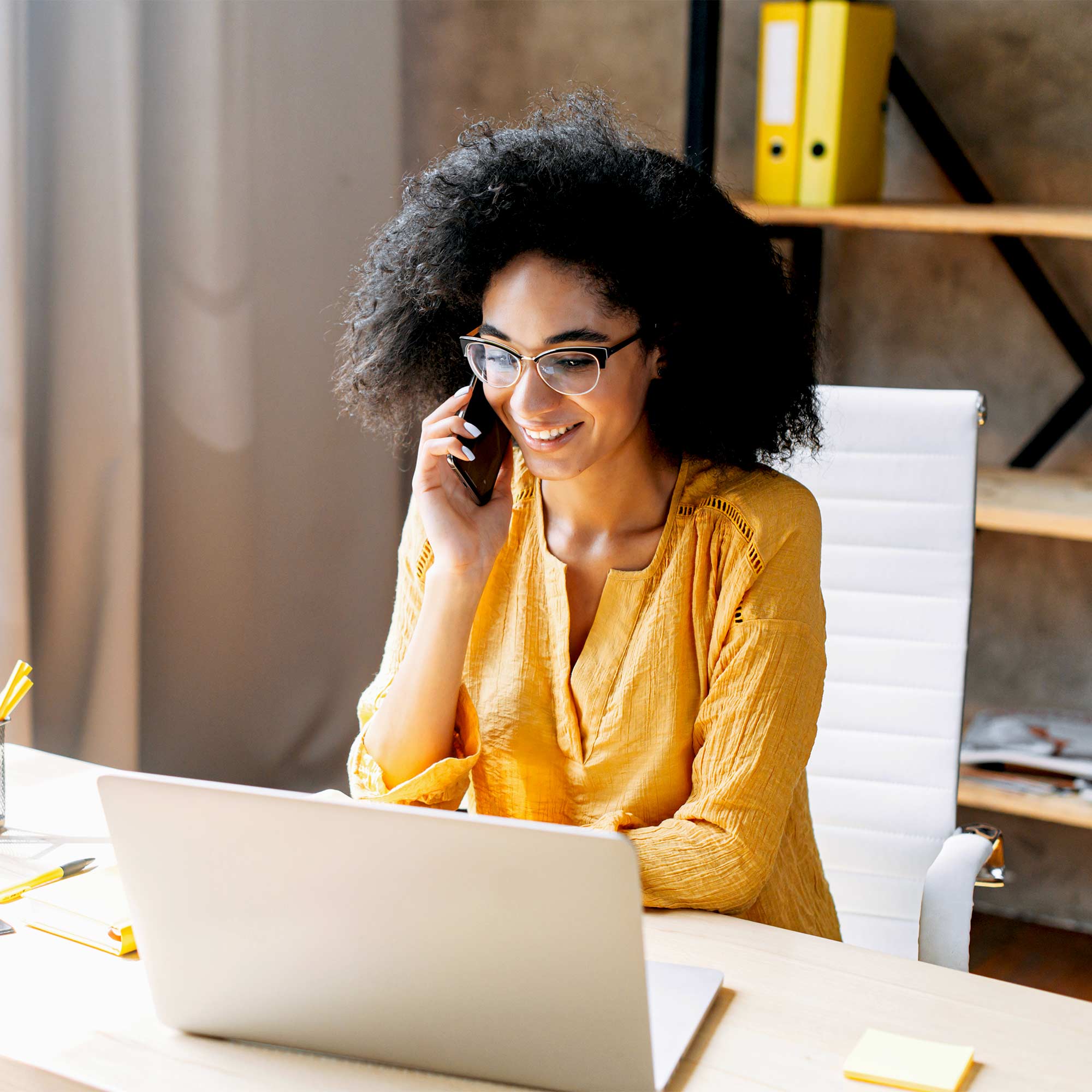 A woman wearing yellow talks on the phone as she sits at her workstation.