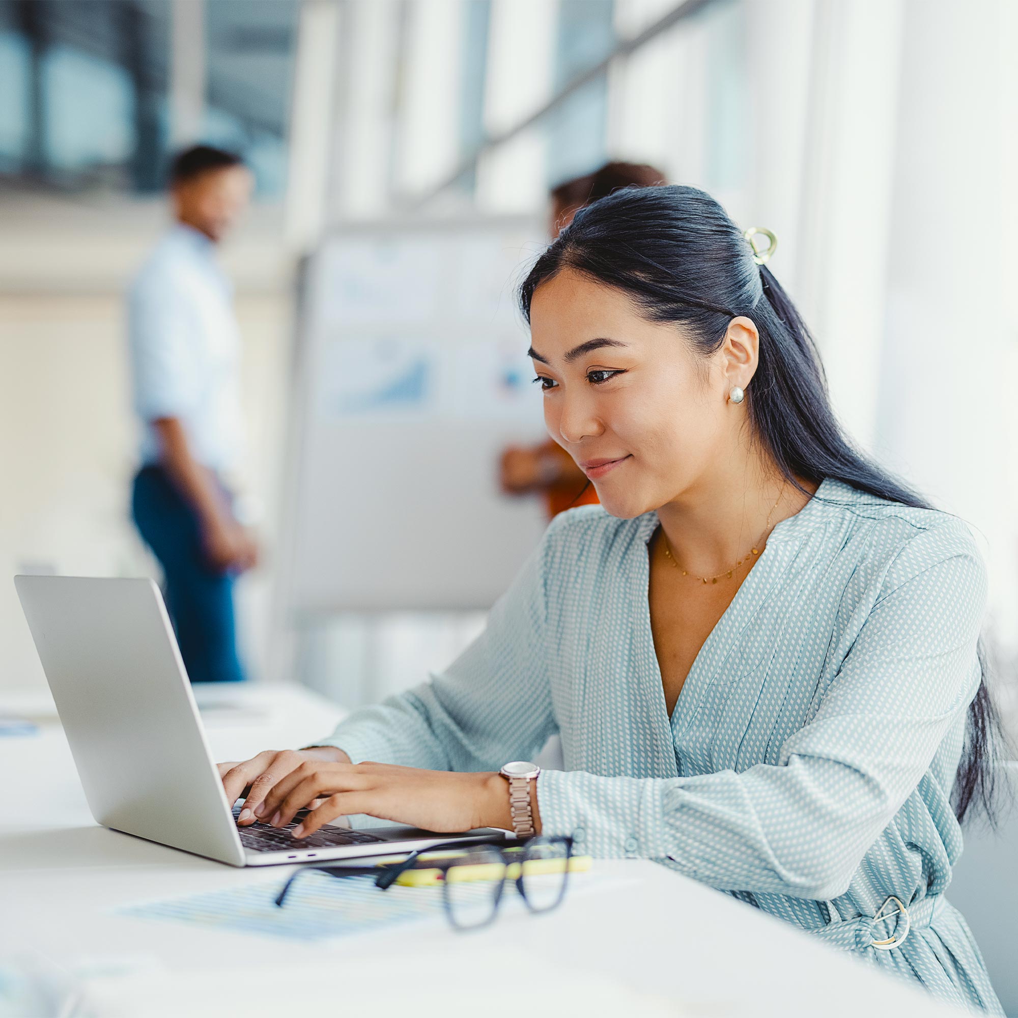 A woman sits and types on her computer as coworkers stand in background.