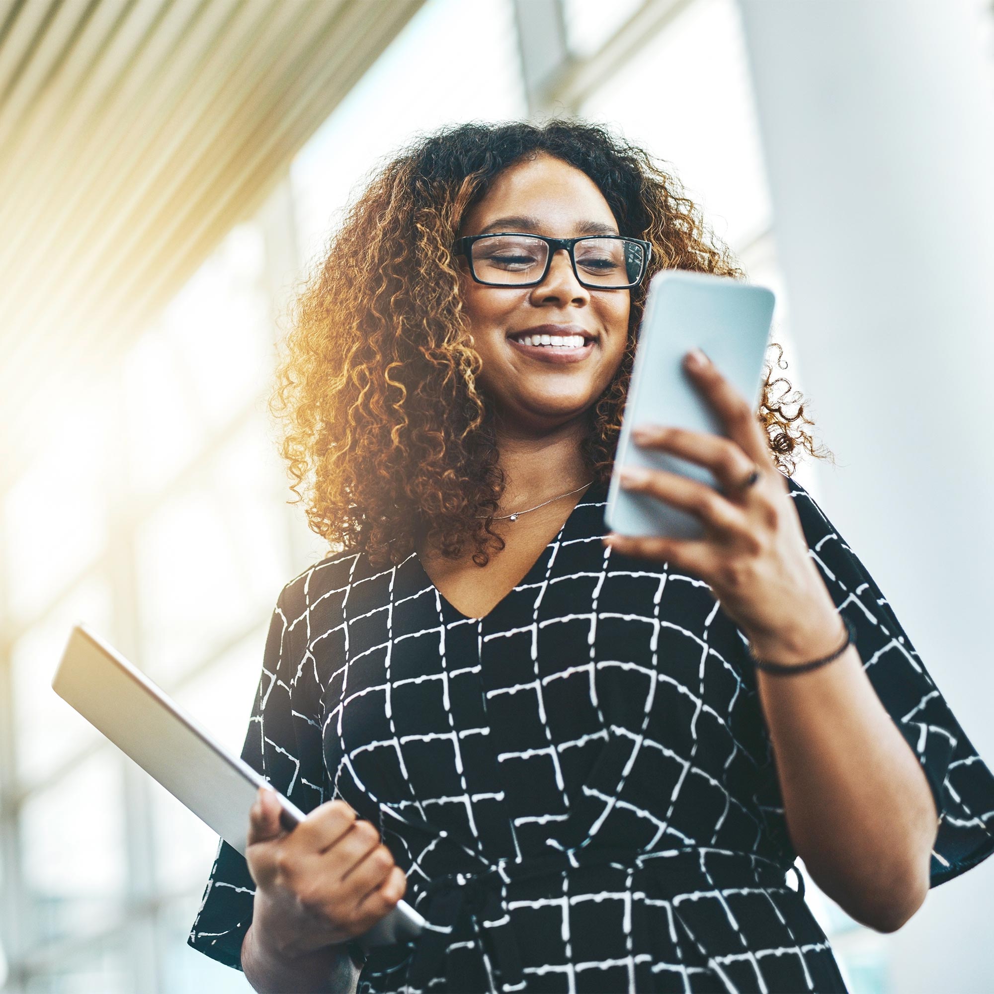 A woman stands and holds her notebook as she smiles at her cell phone.
