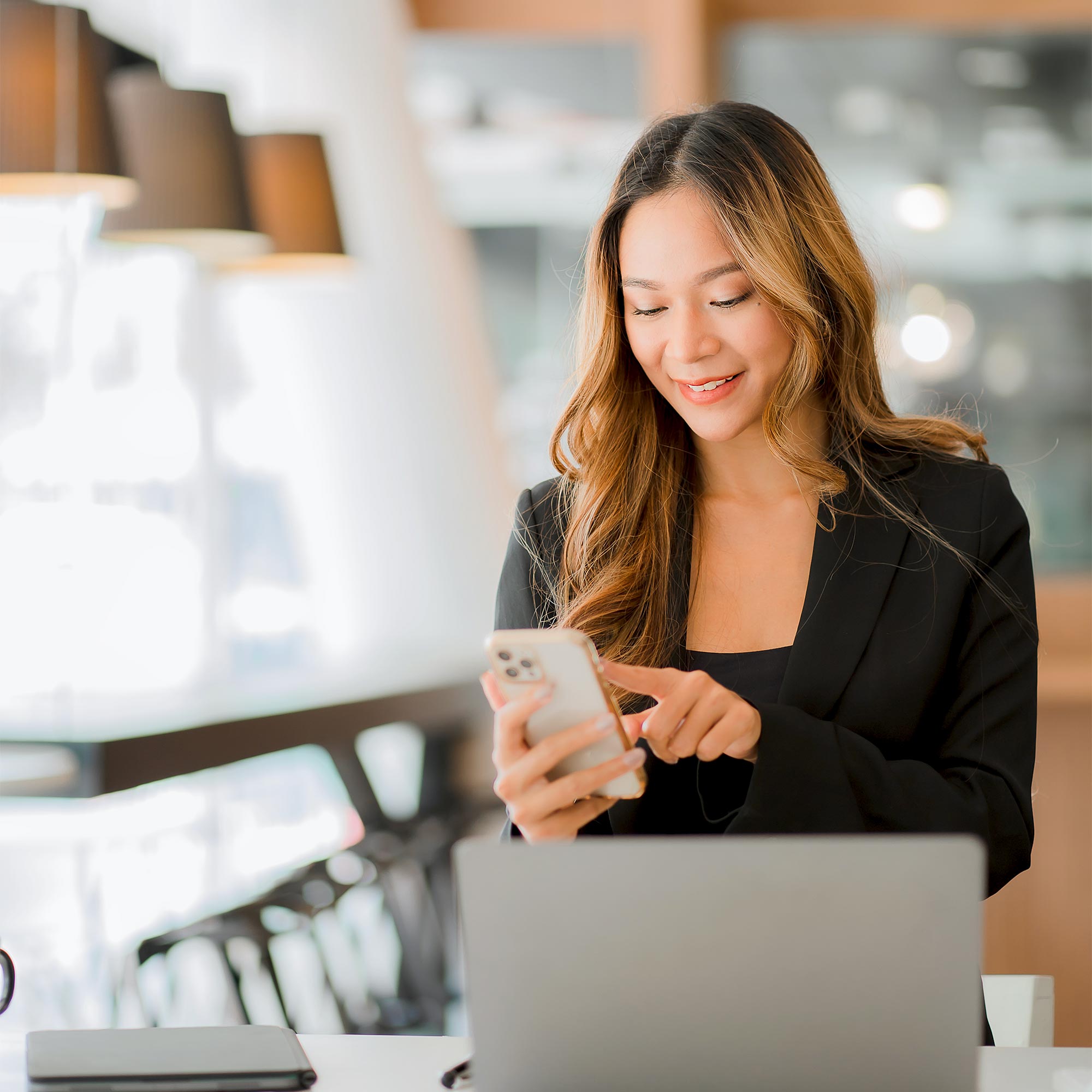 A woman stands near a laptop looking at her cell phone and smiling.
