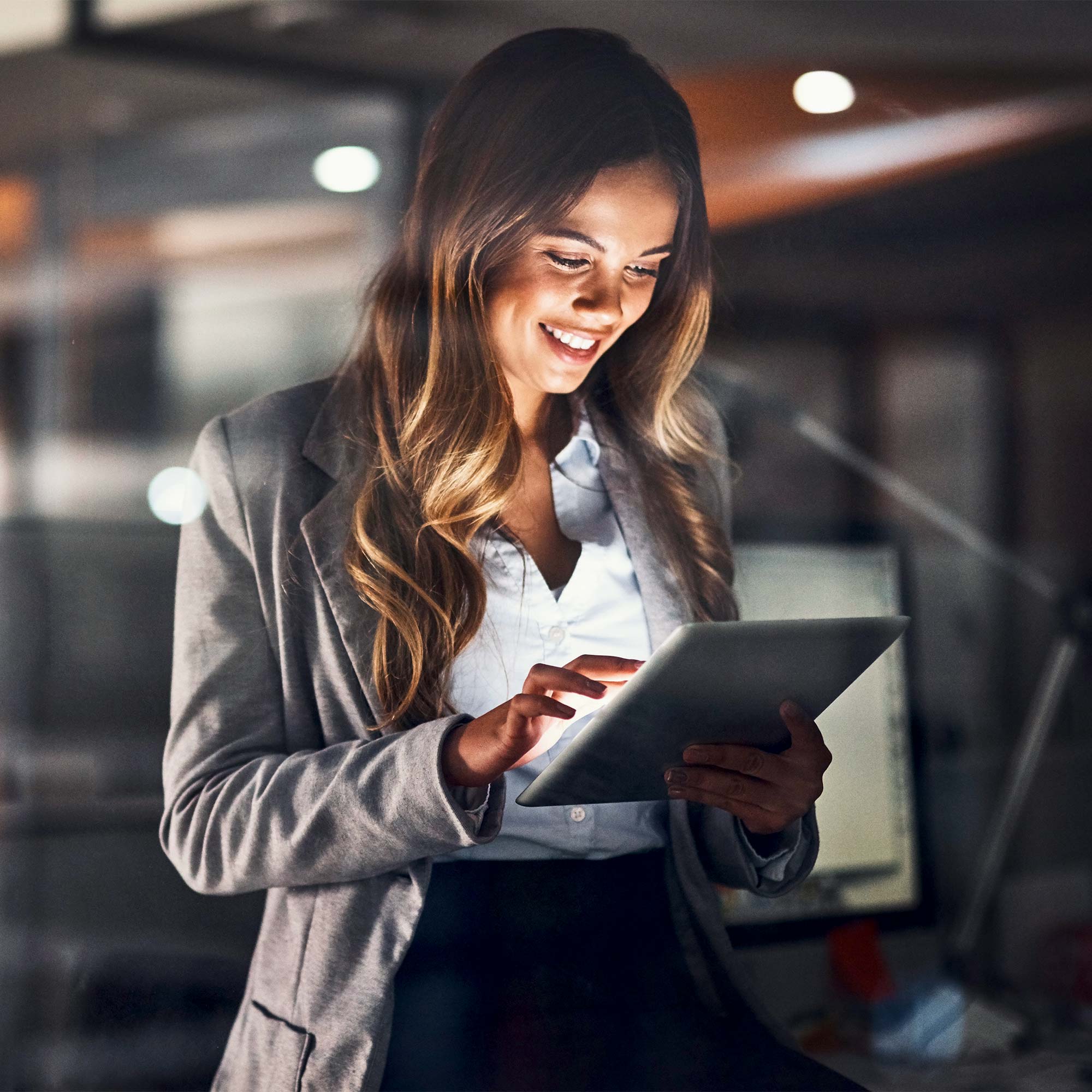 A woman stands near a laptop looking at her cell phone and smiling.