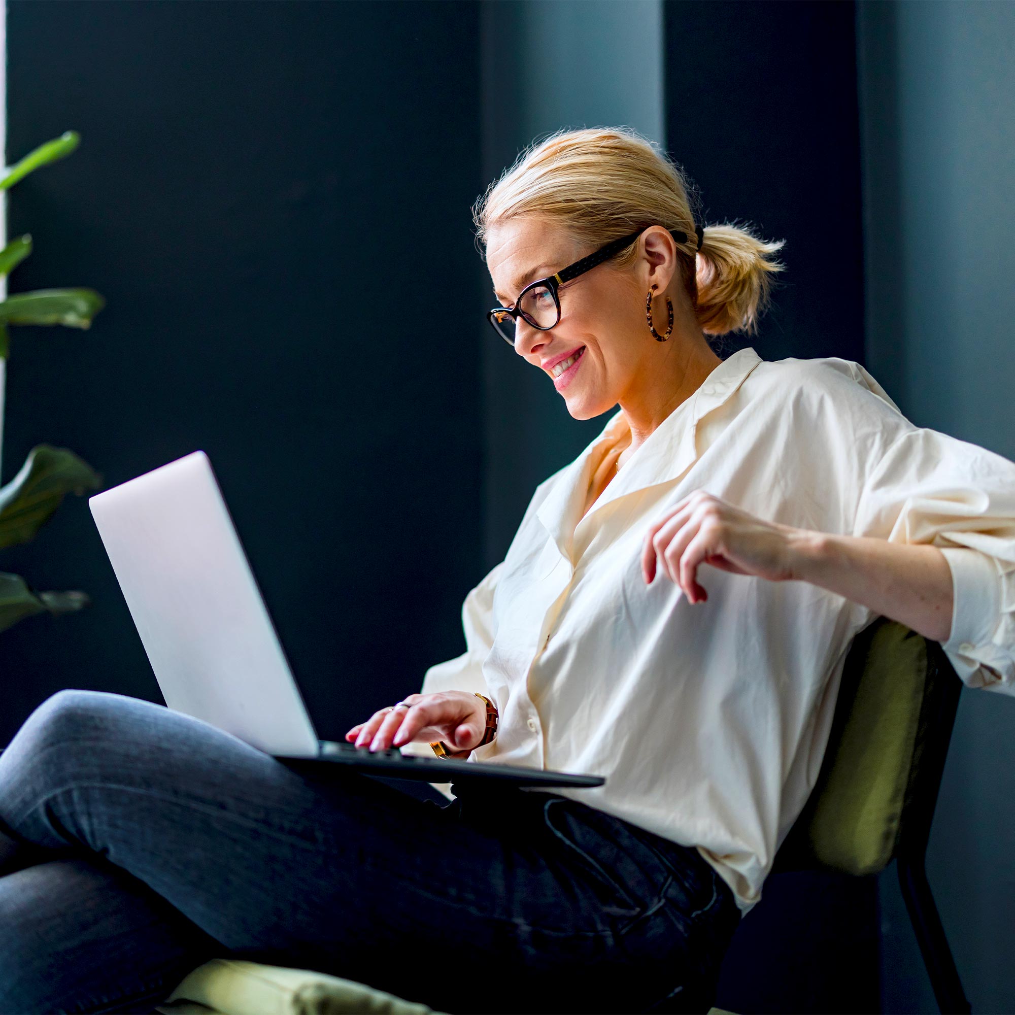 A woman in jeans sits casually and looks at her laptop.