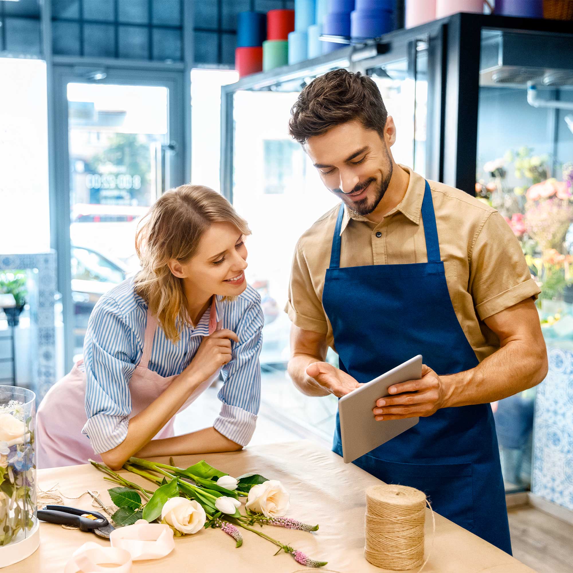 A man and woman wearing aprons, smile and look at a notebook.