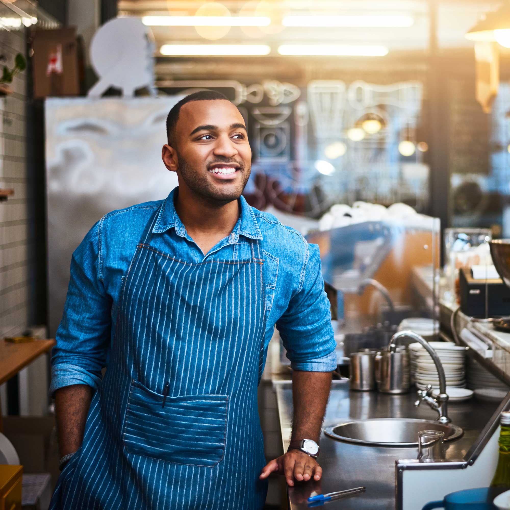 A man in a blue apron smiles as he stands in the kitchen.