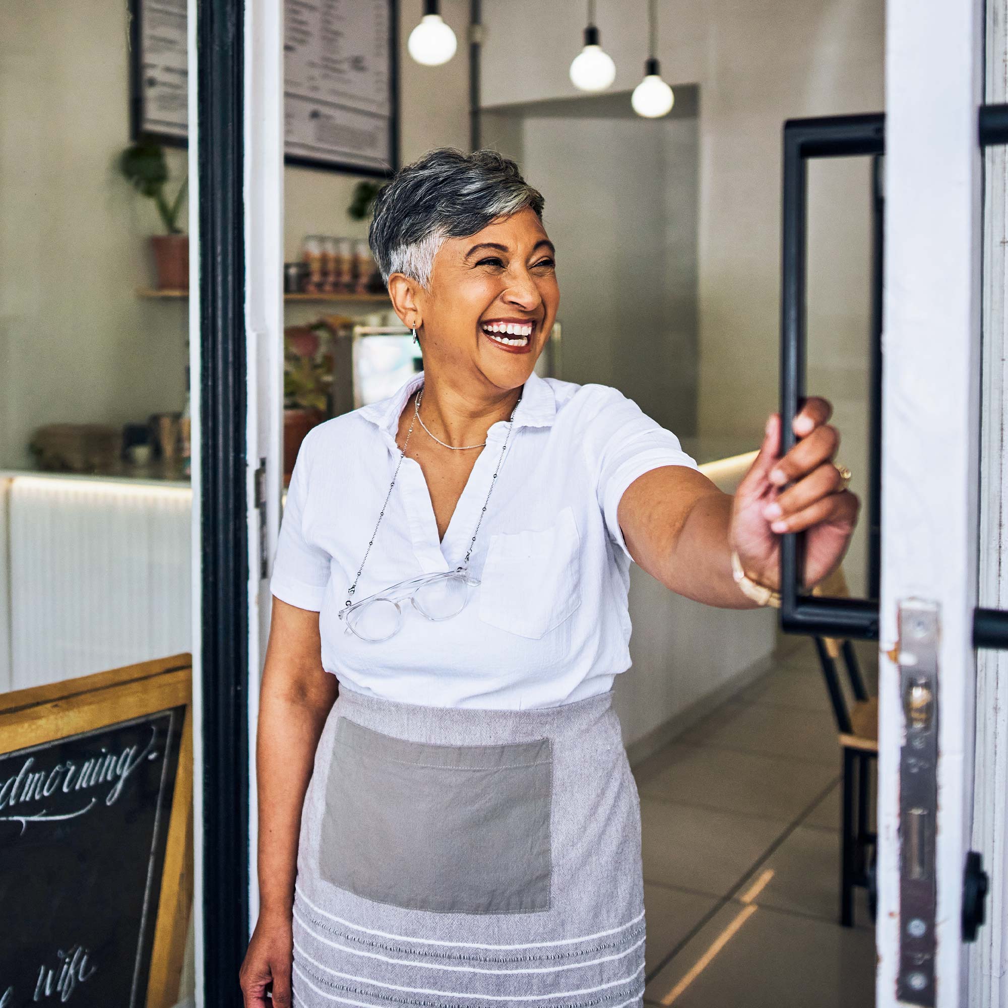 A woman wearing an apron smiles as she holds the door open.