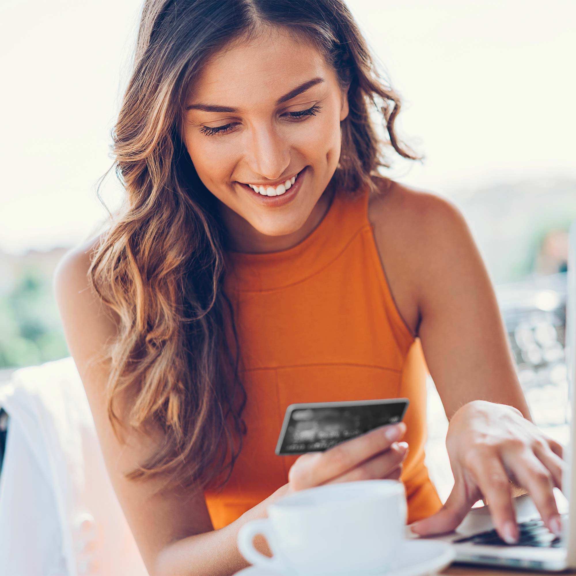 Woman wearing an orange shirt smiling at a credit card while typing on a laptop