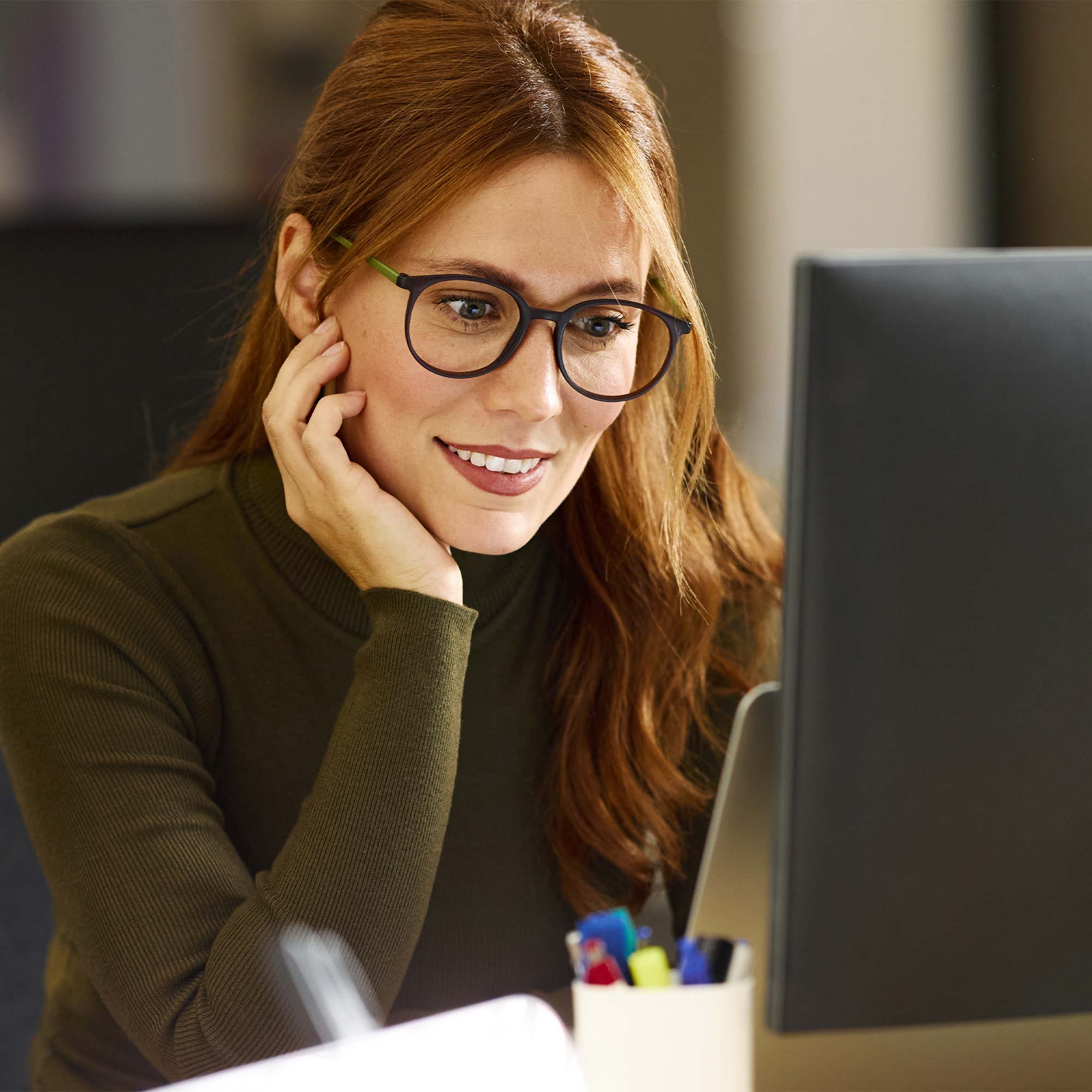 Smiling woman wearing a sweater sitting at a desk with a glass of water, a notebook, and a tablet while using her phone
