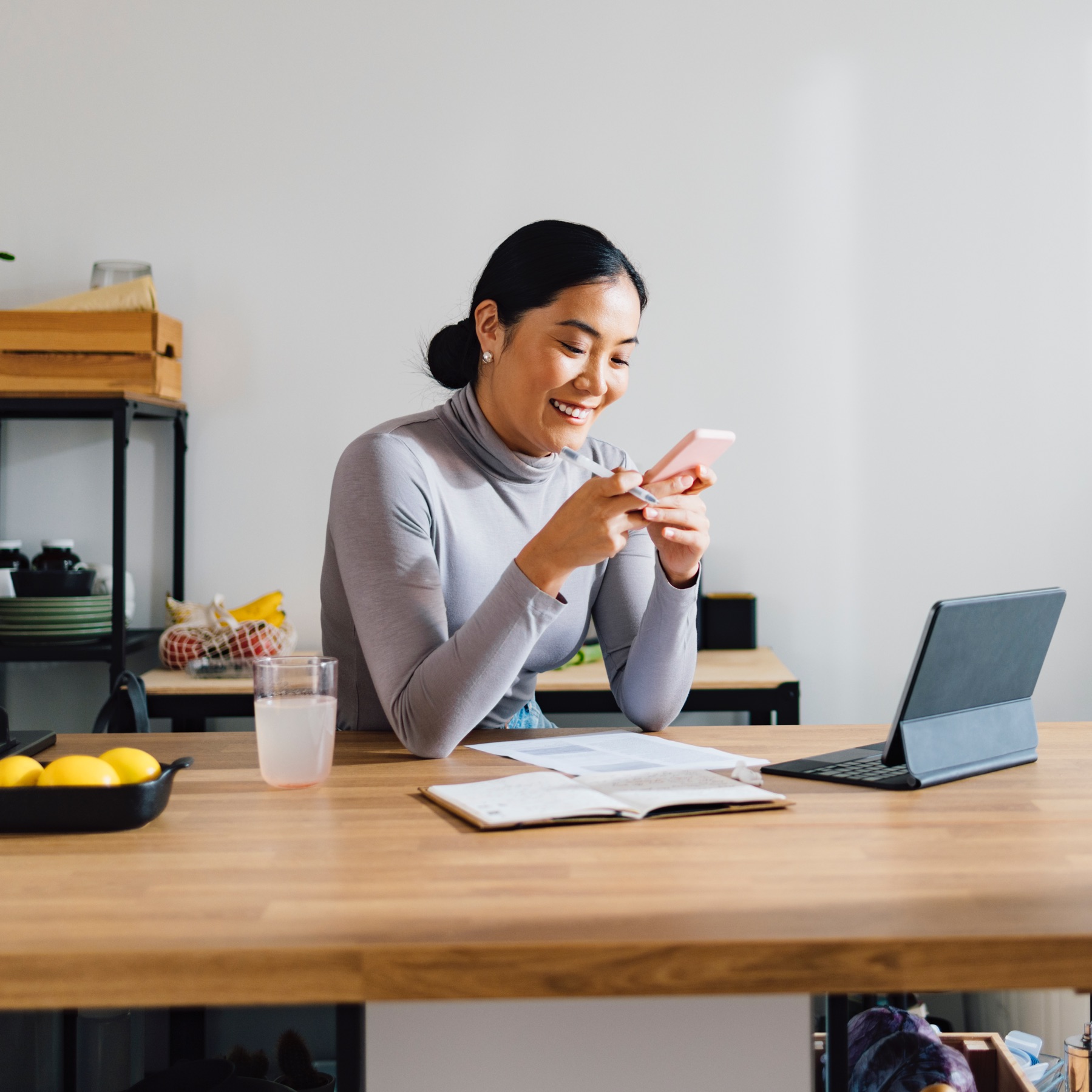 Smiling woman wearing a sweater sitting at a desk with a glass of water, a notebook, and a tablet while using her phone