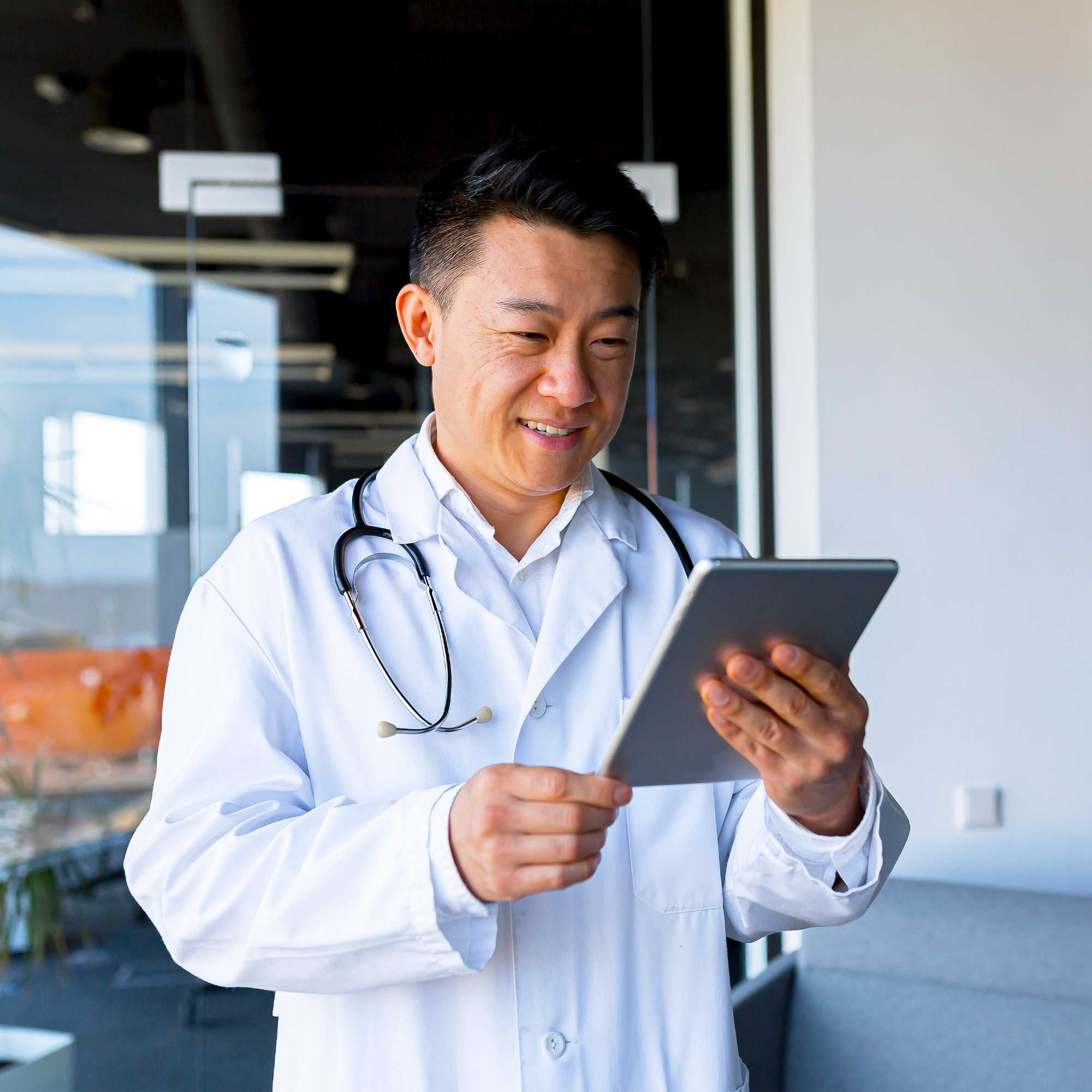 A doctor wearing a stethoscope, smiles as he looks a tablet.