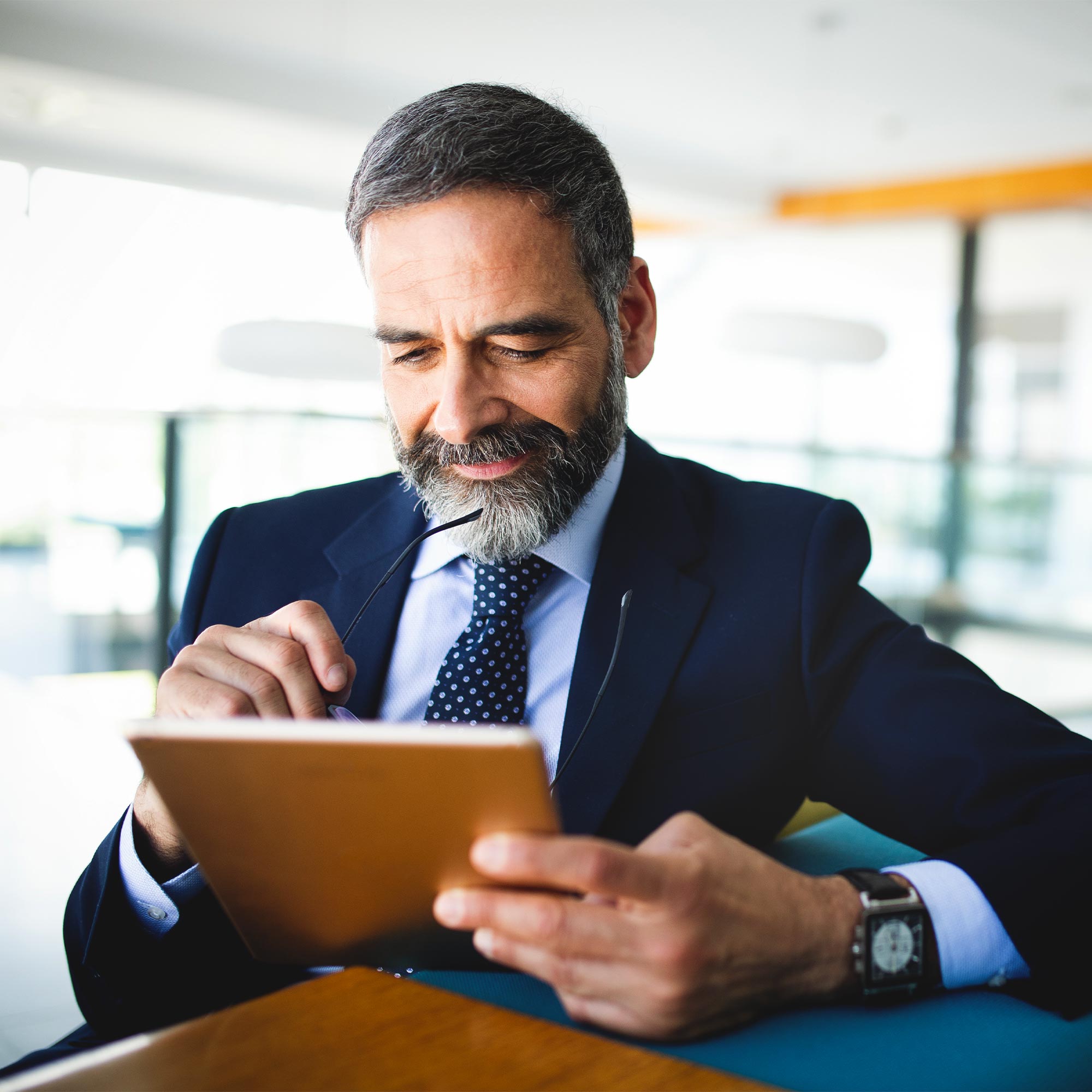 Two professionally dressed men sit at a gray table and look at a tablet.