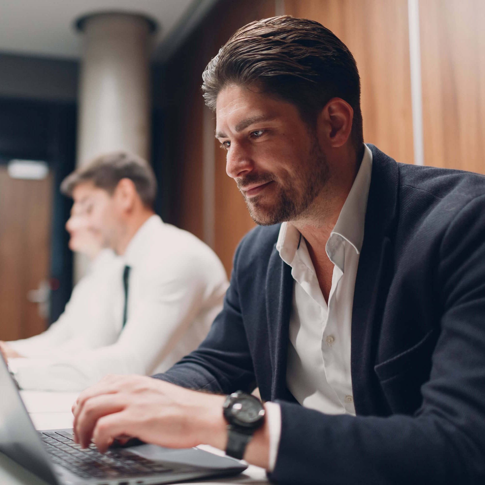 A professionally dressed man sits at a table working on a laptop.