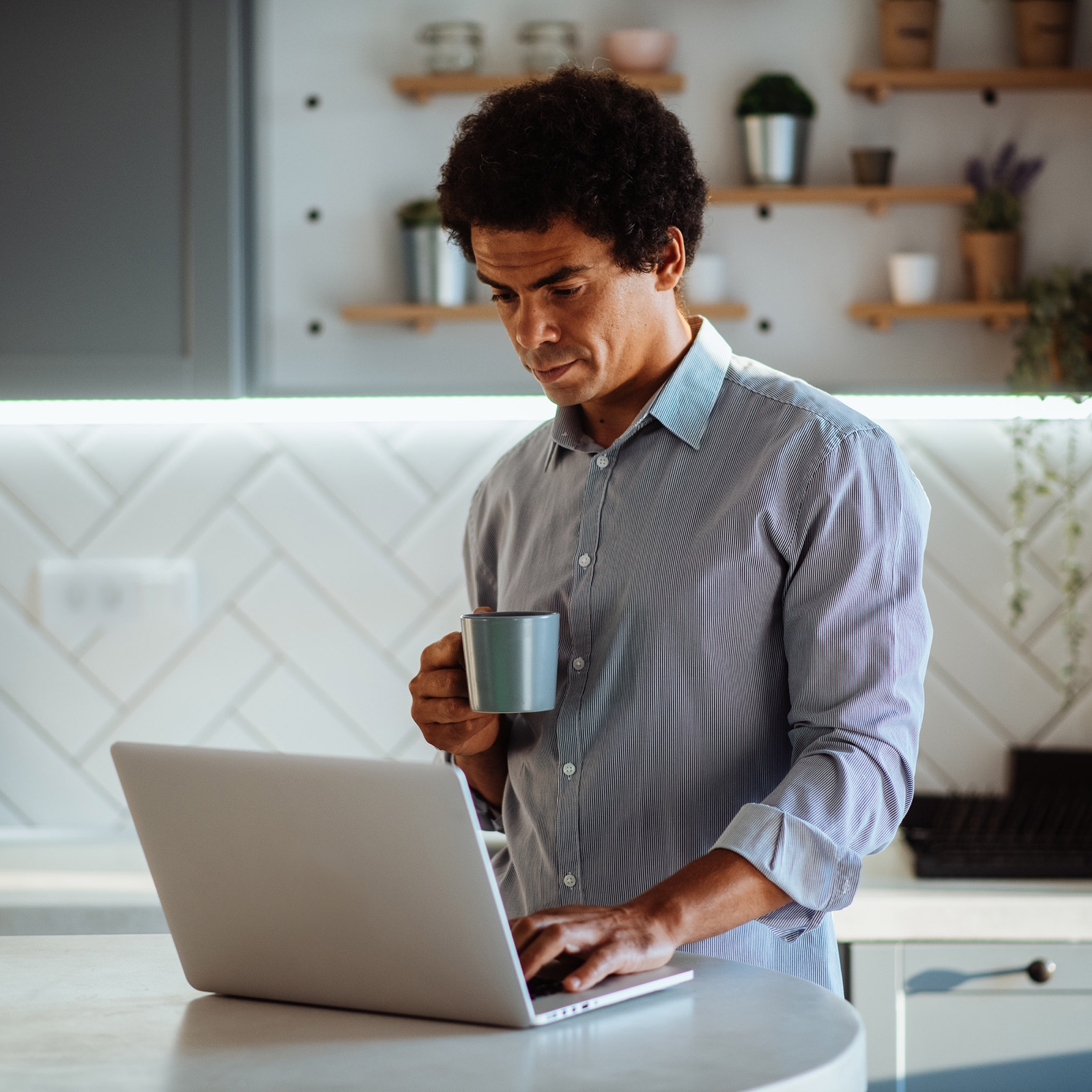 Man in a blue and white striped button up shirt holding a cup of coffee and typing something on his laptop