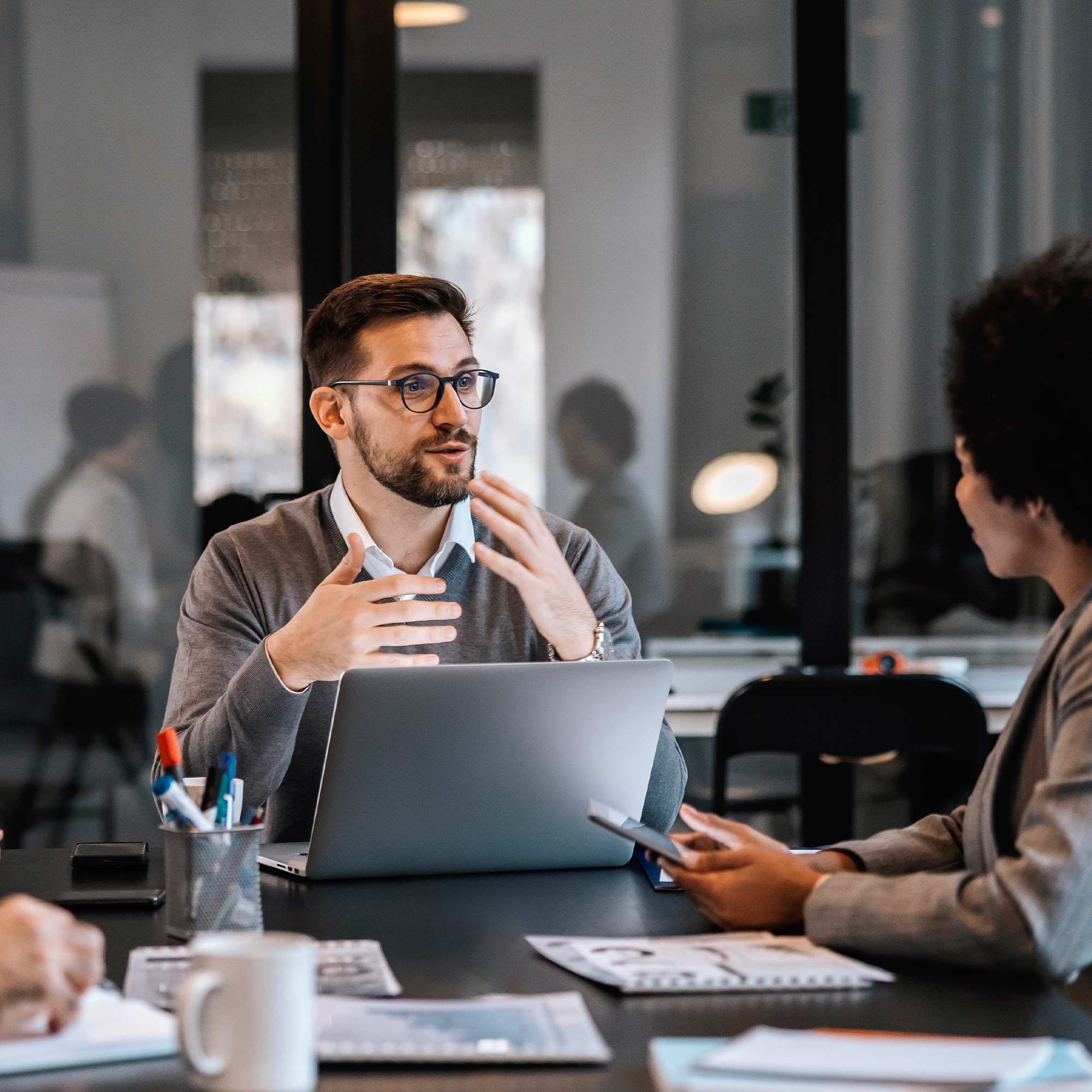 A man in a business suit sits at a desk with a cup of coffee and a coworker.