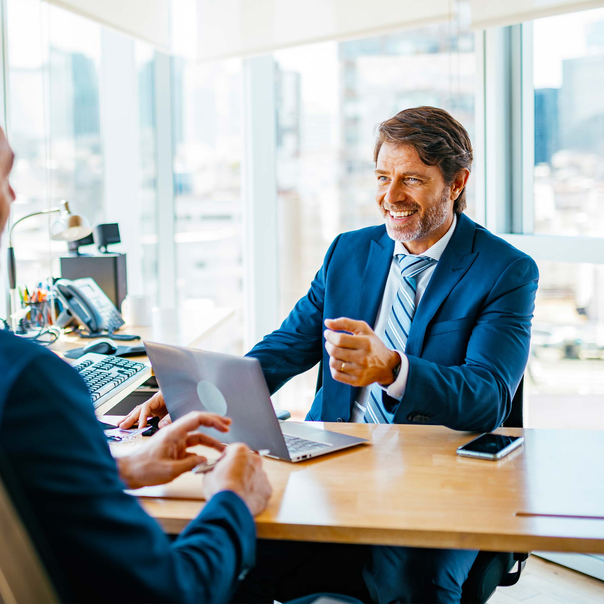 A man in a business suit sits at a desk with a cup of coffee and a coworker.