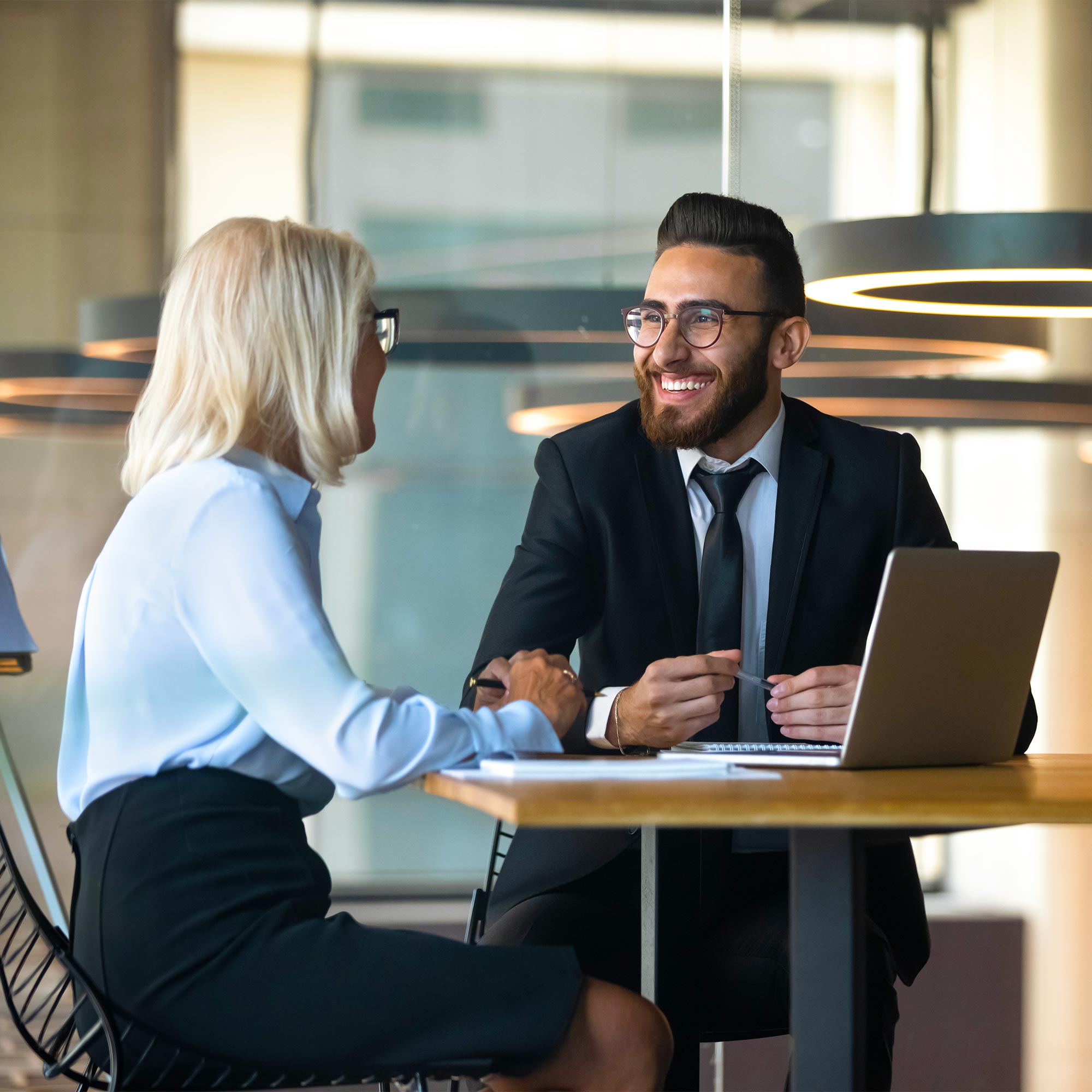 A professionally dressed man sits across the table from a woman and smiles.