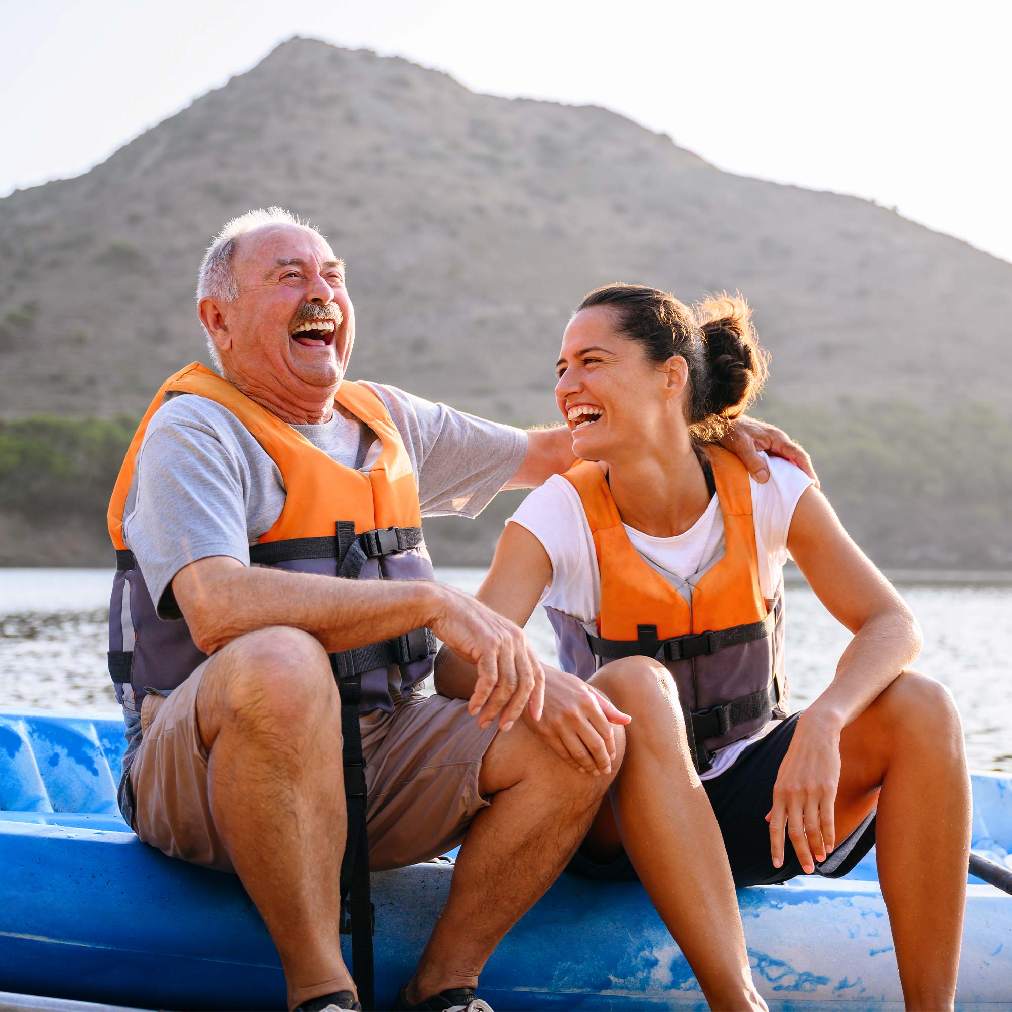 A father and daughter wearing life jackets sit on a raft laughing.
