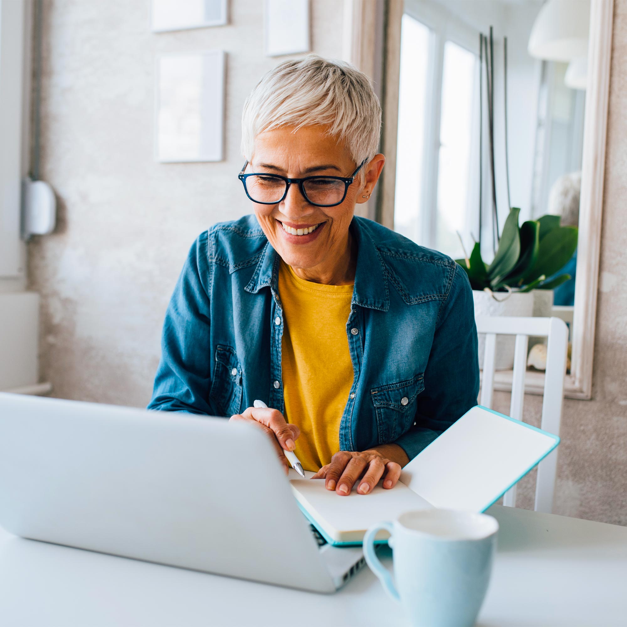 A woman wearing a yellow shirt and blue jacket uses her laptop and writes in her notebook.
