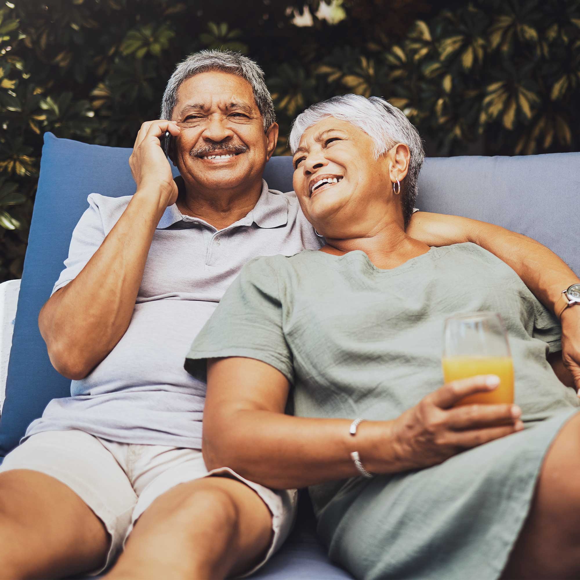 A man and woman sit on a patio chair smiling as he talks on phone.