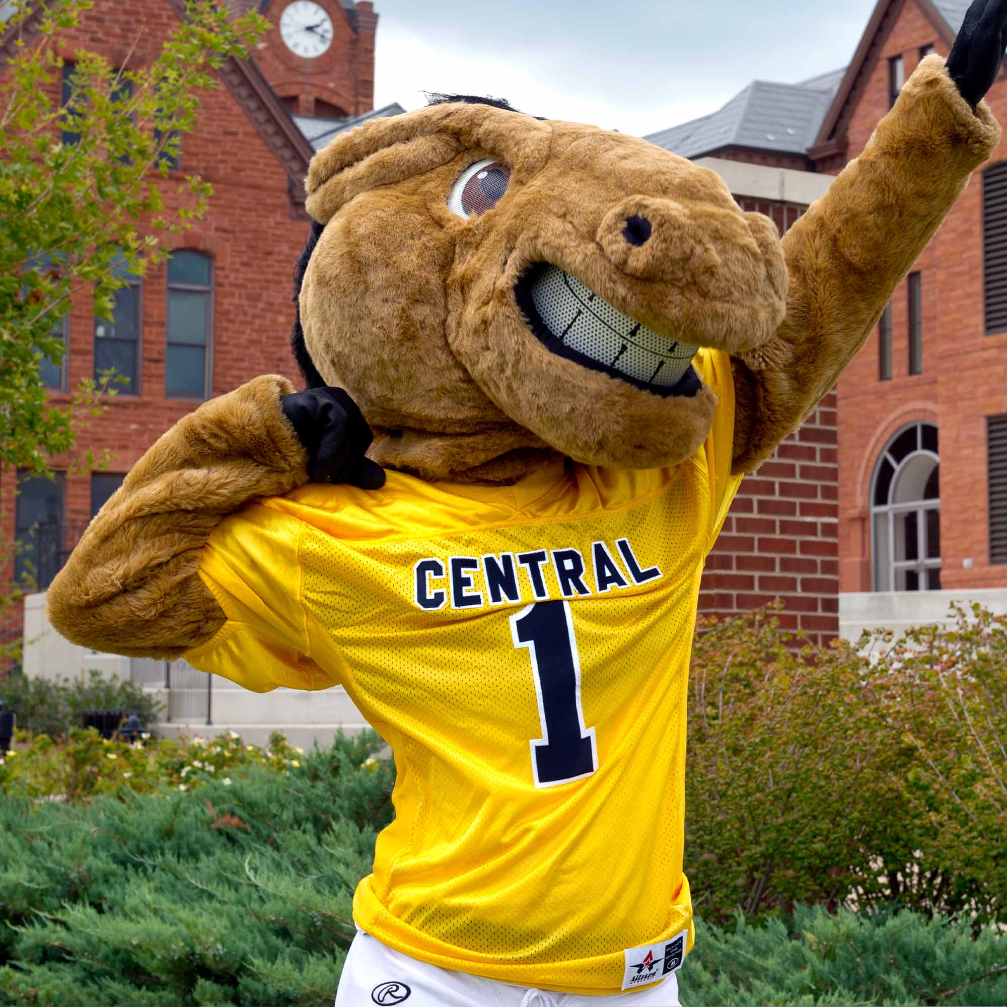 Mascot Buddy the Broncho raises his hands toward the sky in front of the UCO clock tower.