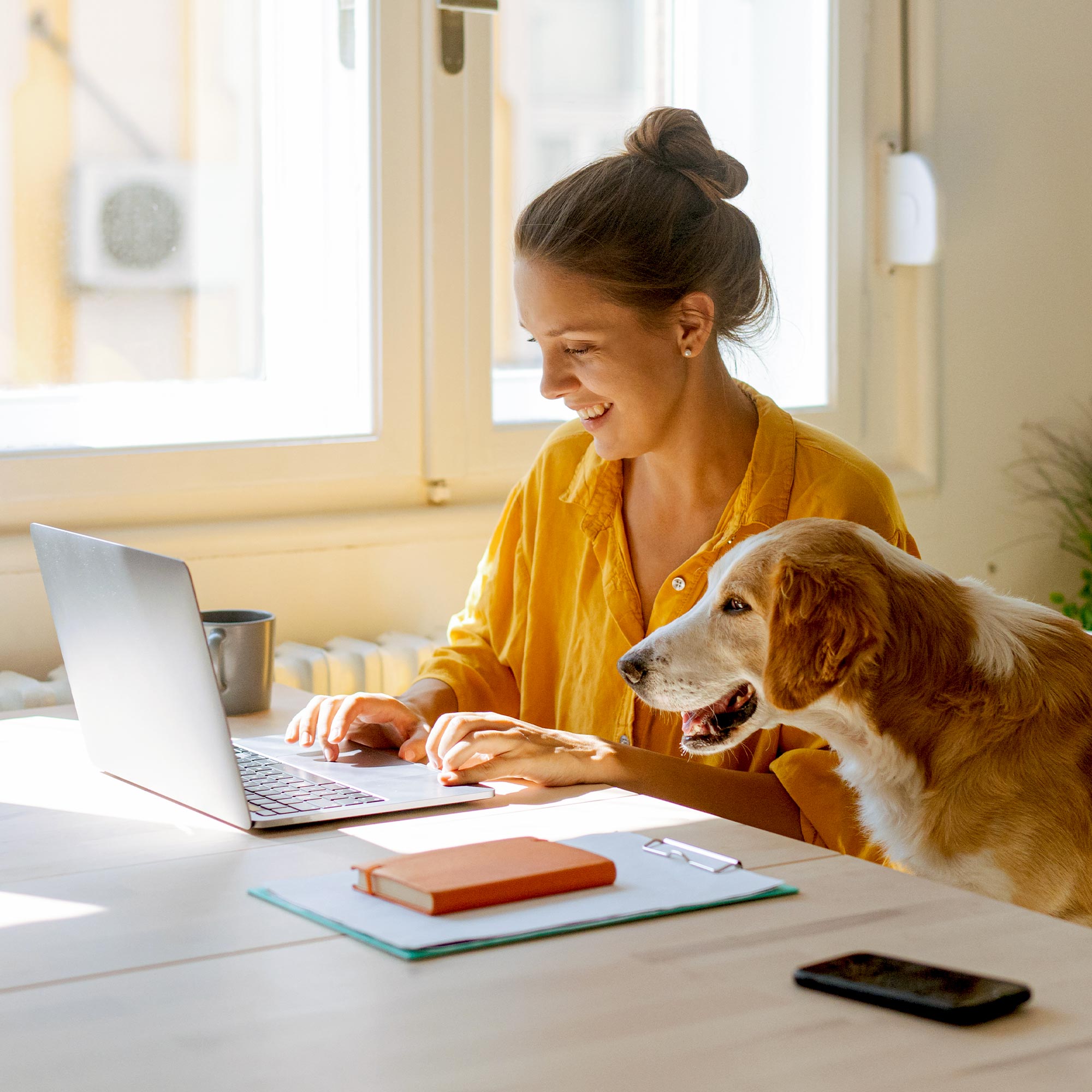 A woman in a yellow shirt uses a computer with her dog standing next to her.