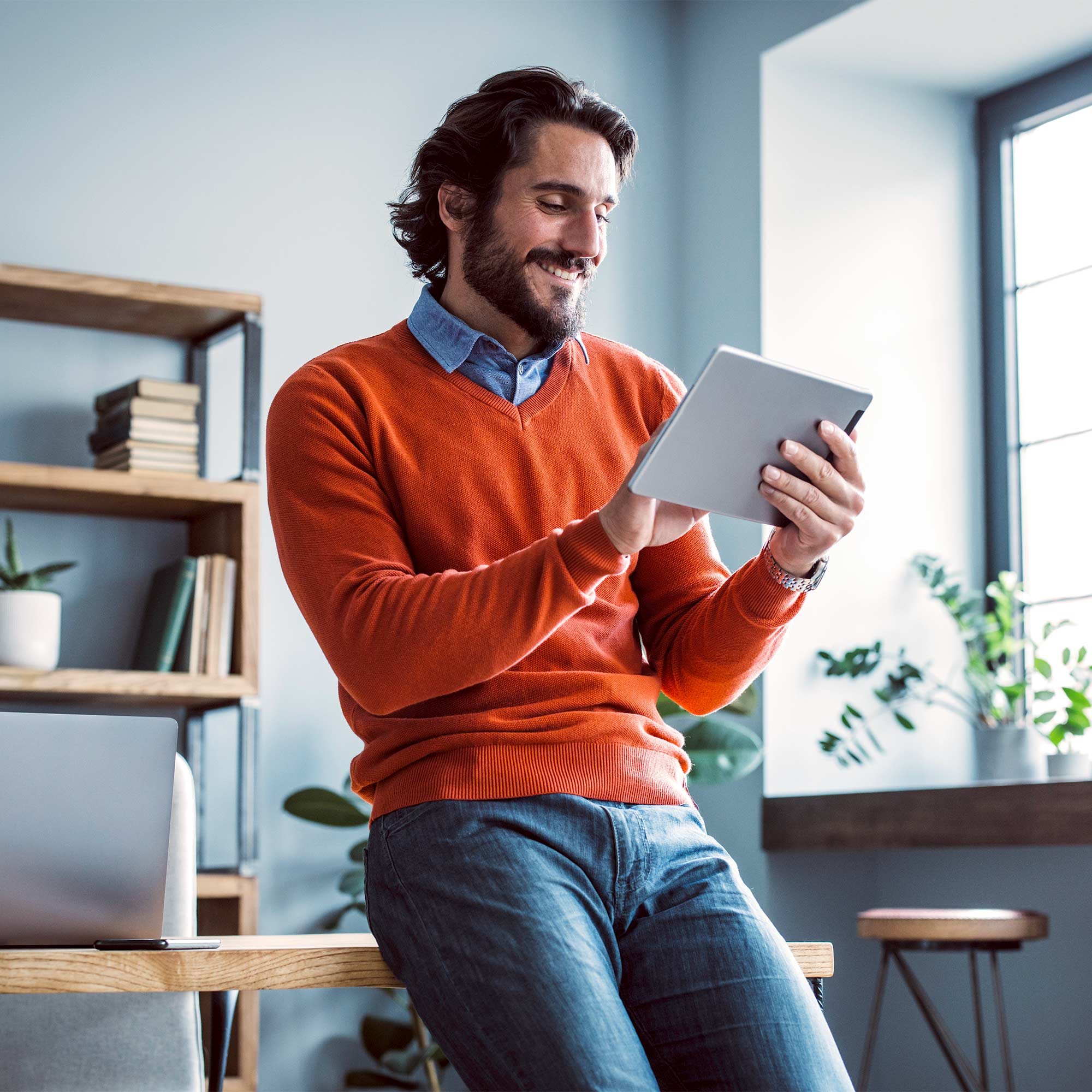A man wearing an orange sweater smiles as he uses his tablet.
