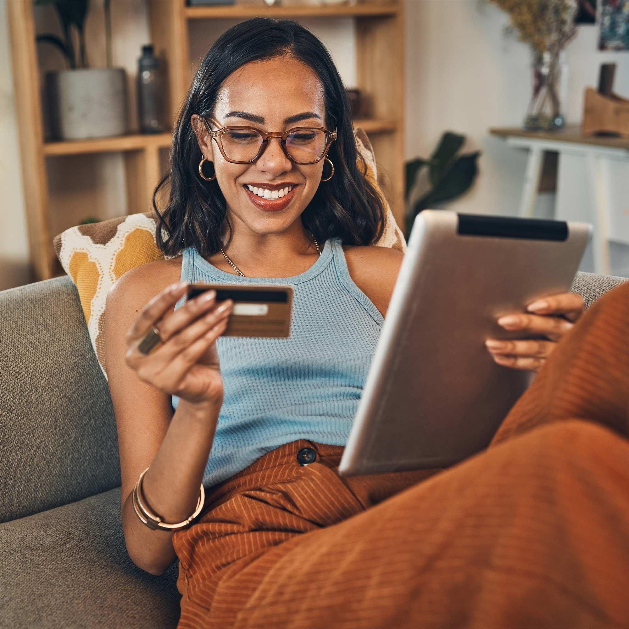 Woman looking and smiling at her debit card and ipad as she reclines on the sofa.
