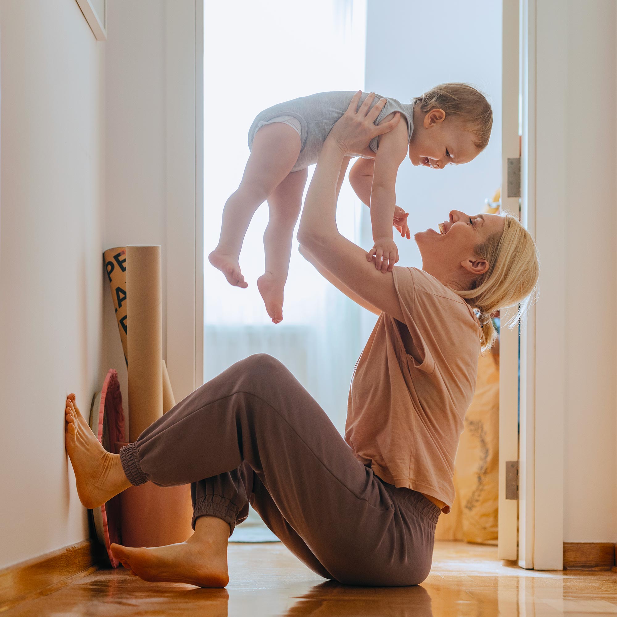 A woman sits on the floor as she holds her baby in the air smiling.