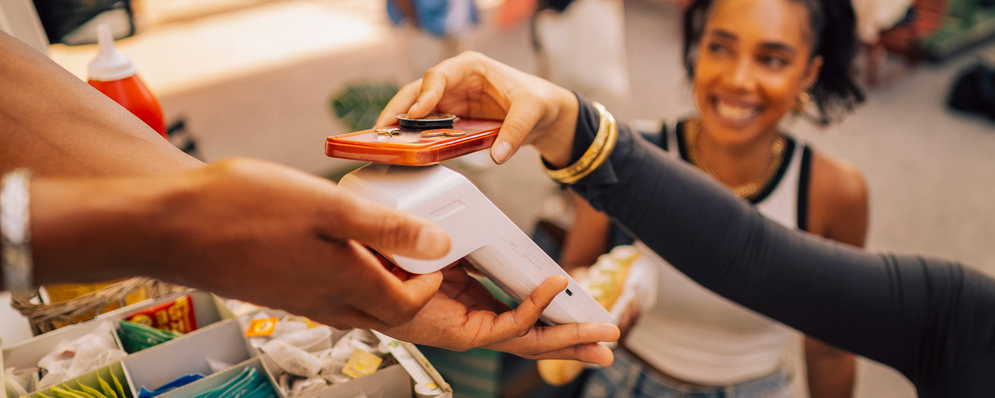 A young woman sits in a kitchen smiling and holding her debit card out to pay.