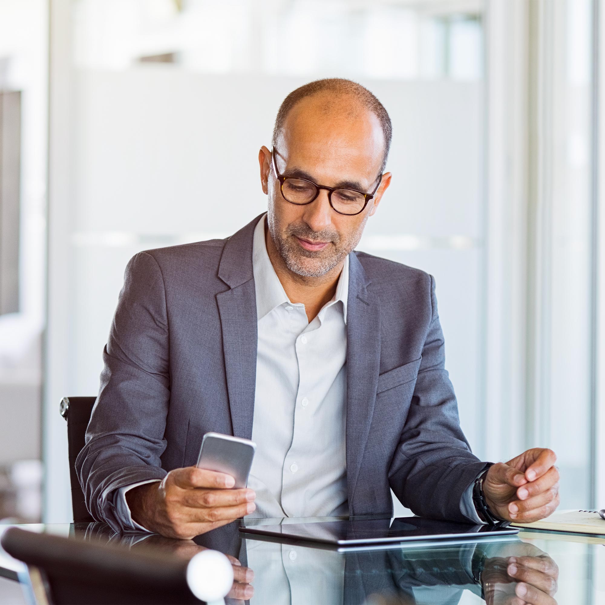 A man wearing a gray suit and glasses looks at his mobile phone as he sits at the desk.