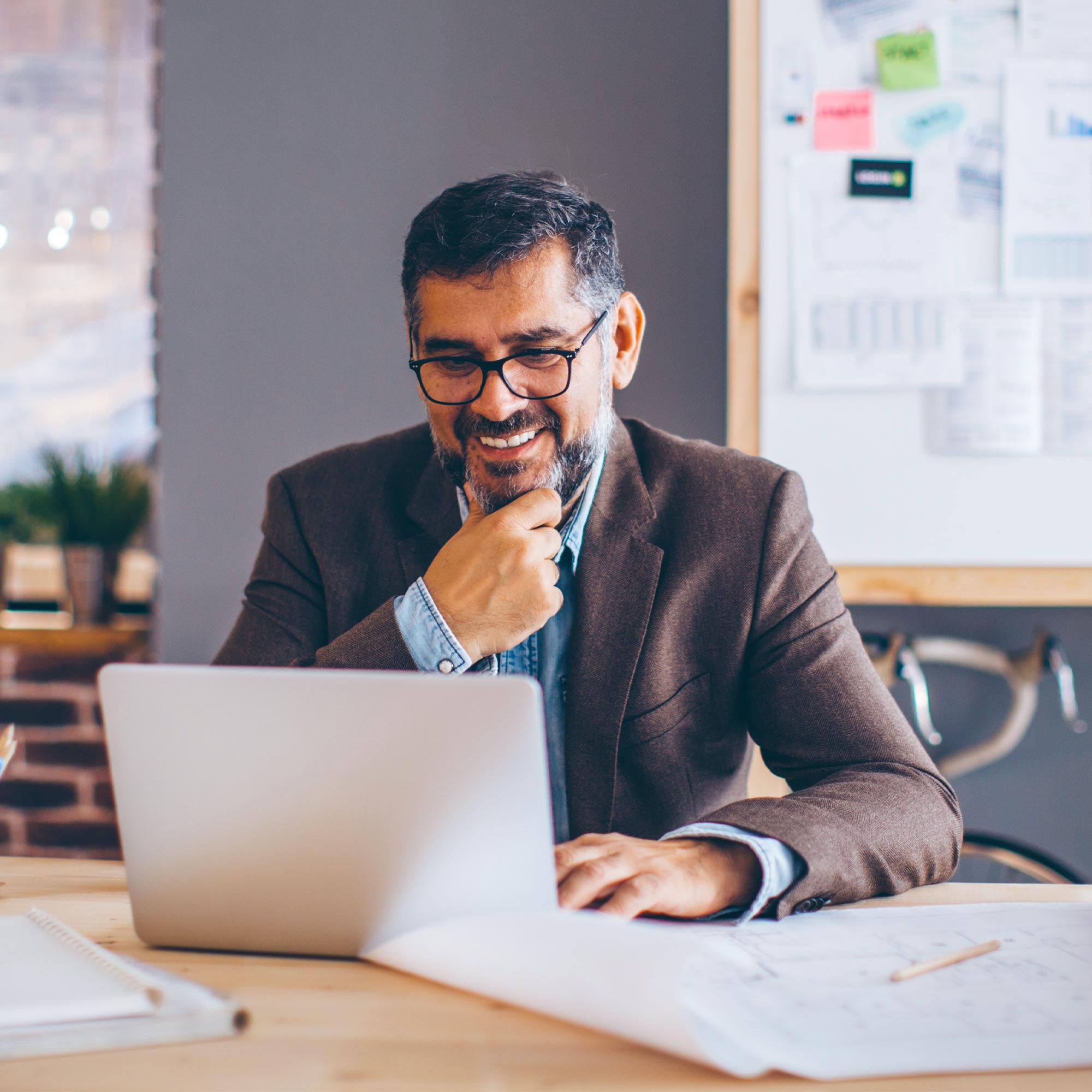 A man wearing a brown jacket and glasses smiles as he looks at his laptop.