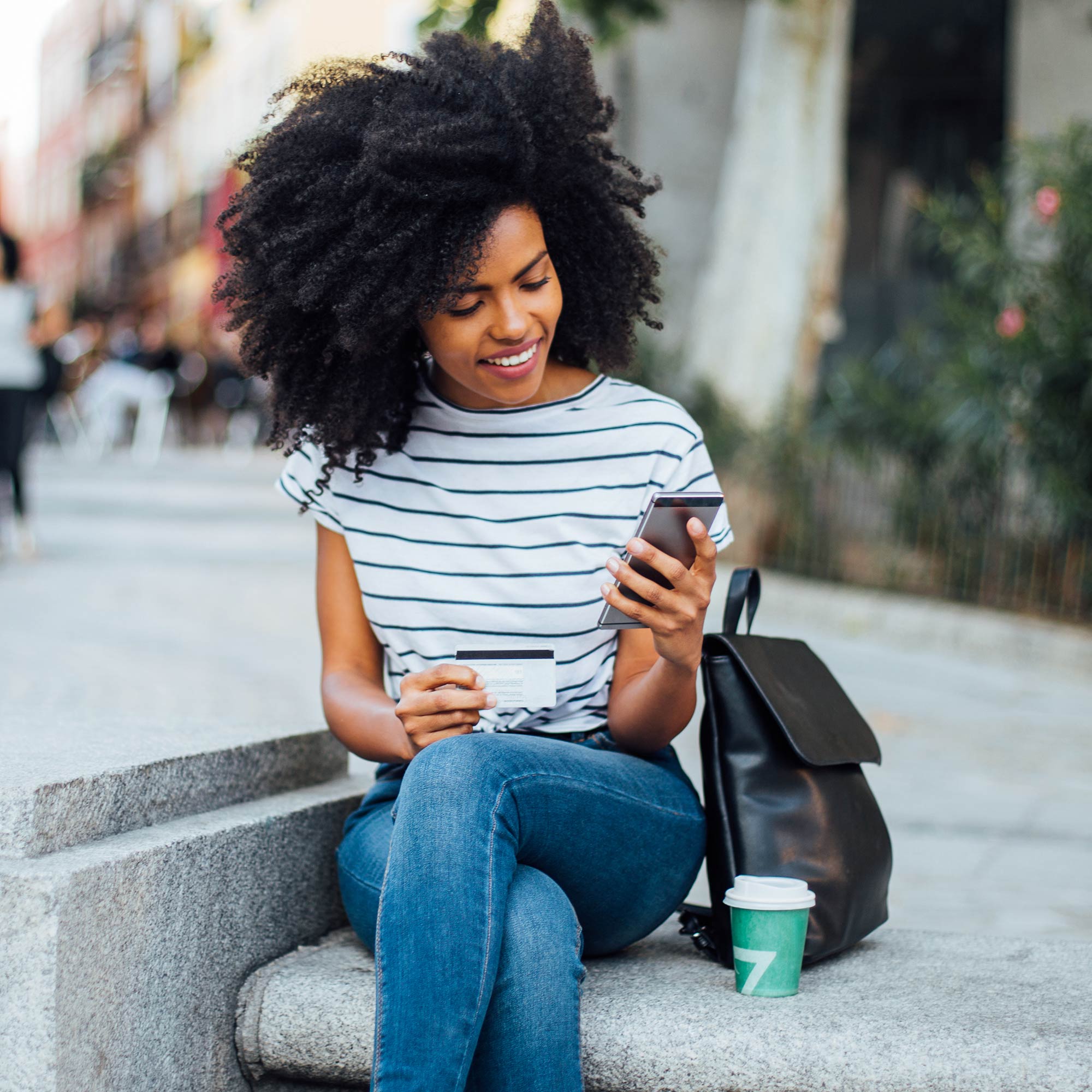 A woman sits on the sidewalk holding her debit card and phone.