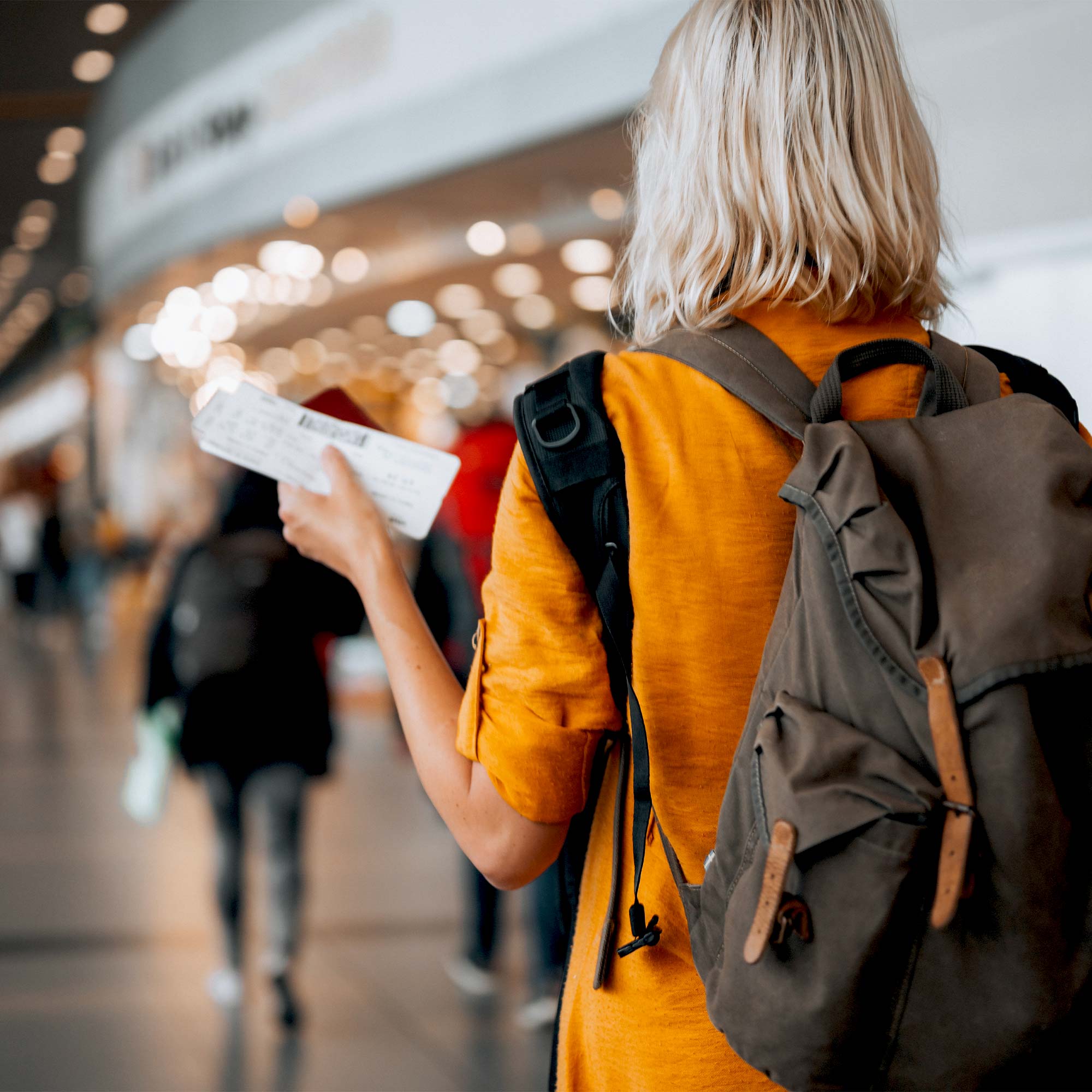 A woman in an orange shirt walks through an airport holding her plane tickets.