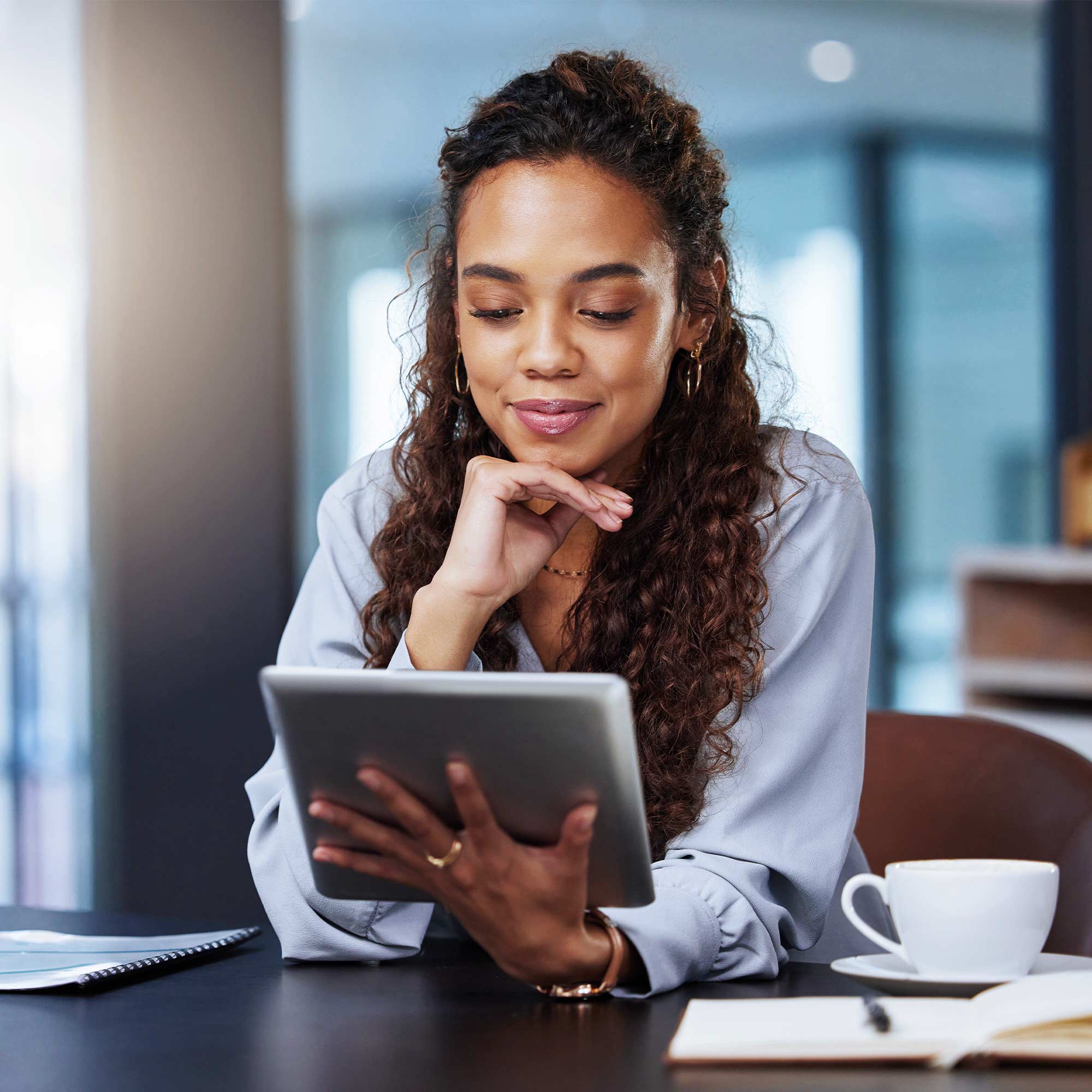 A woman sits at the table with a cup of coffee looking at her tablet.