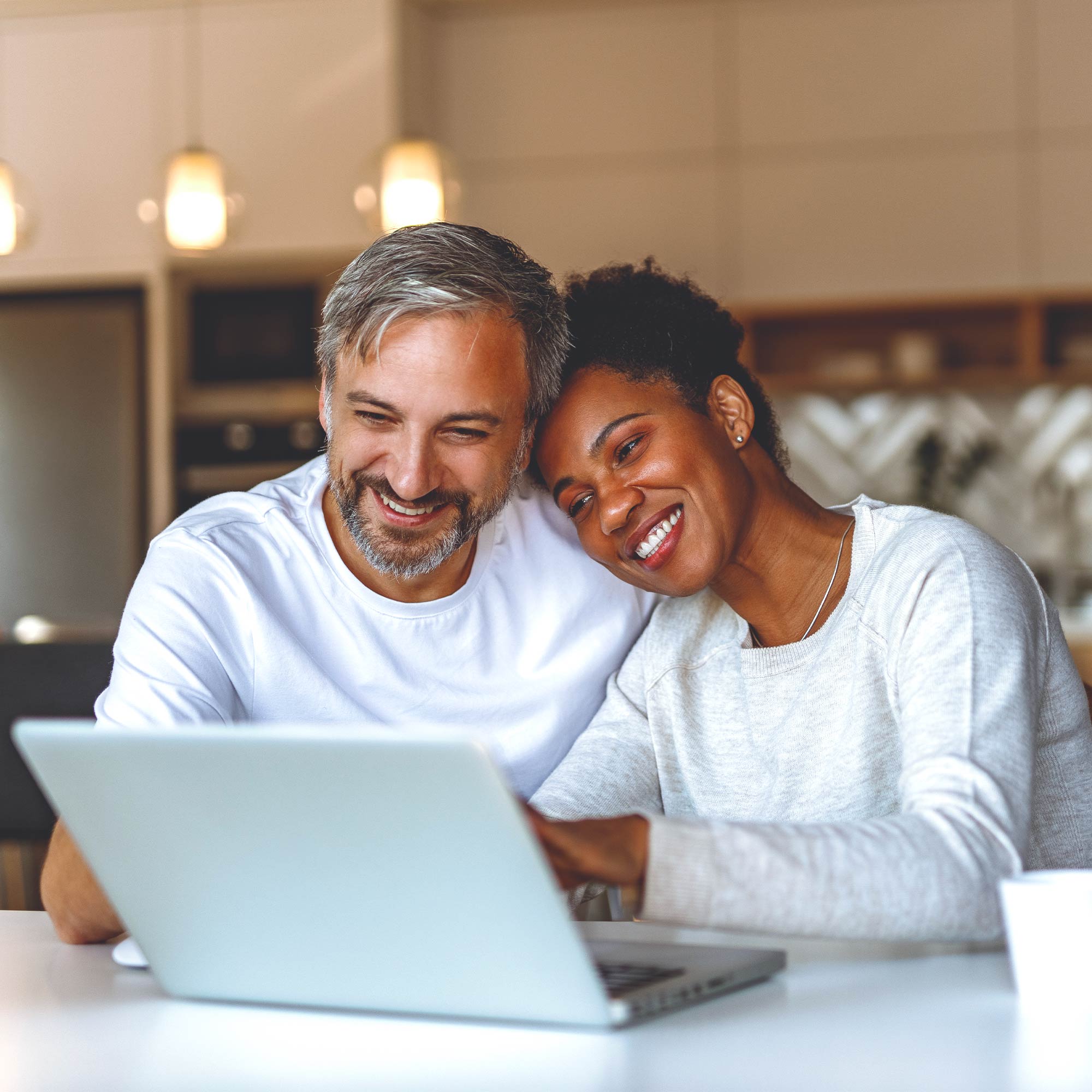 A man and a woman look at their laptop together and smile in their kitchen.