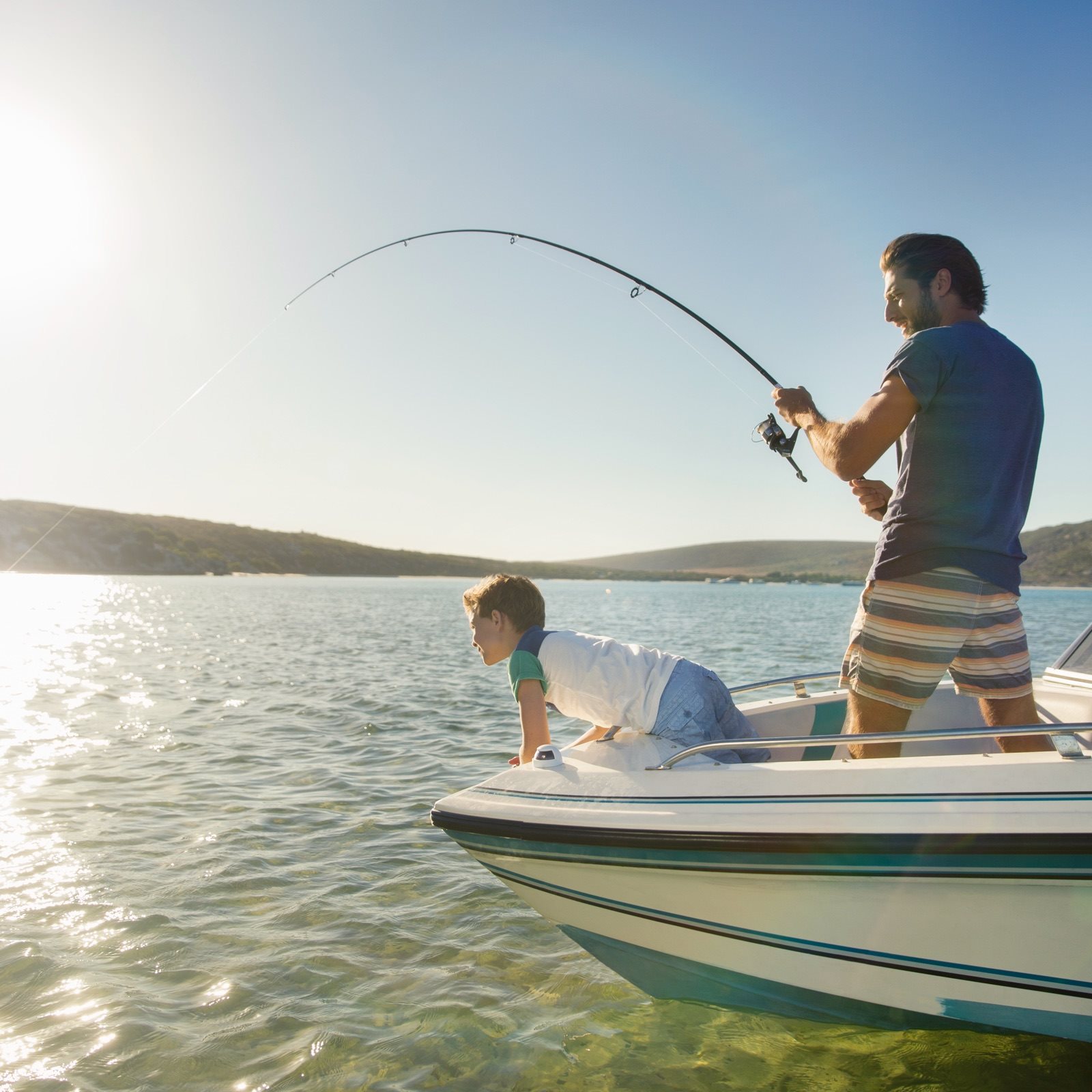A man stands in a boat reeling in a fish while his child leans over and watches.