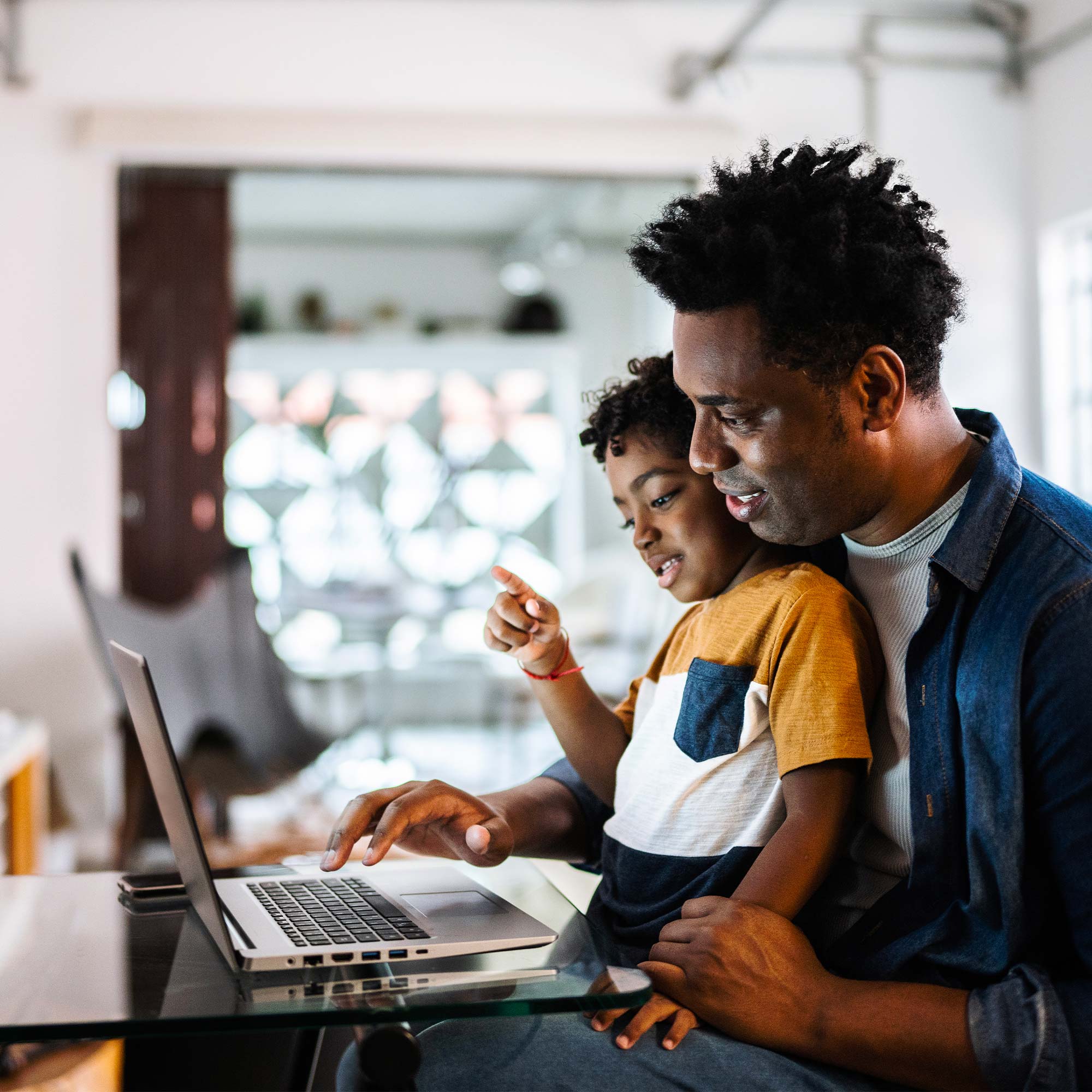 A child sits on her dad's lap as they both look at the computer.