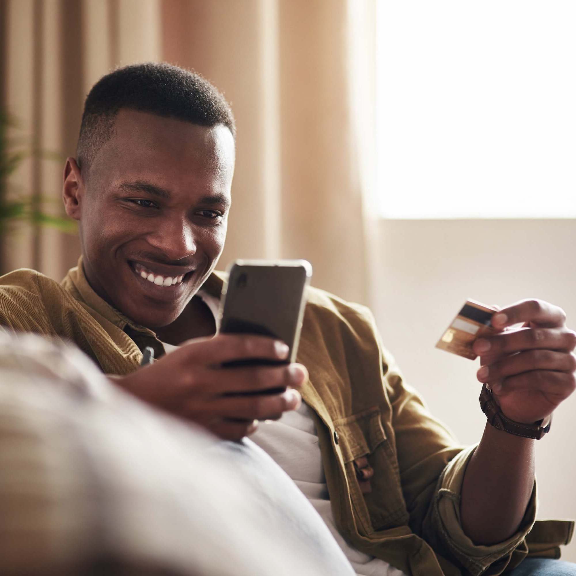 Man wearing a jacket sits on a couch and looks at his phone while holding a debit card.