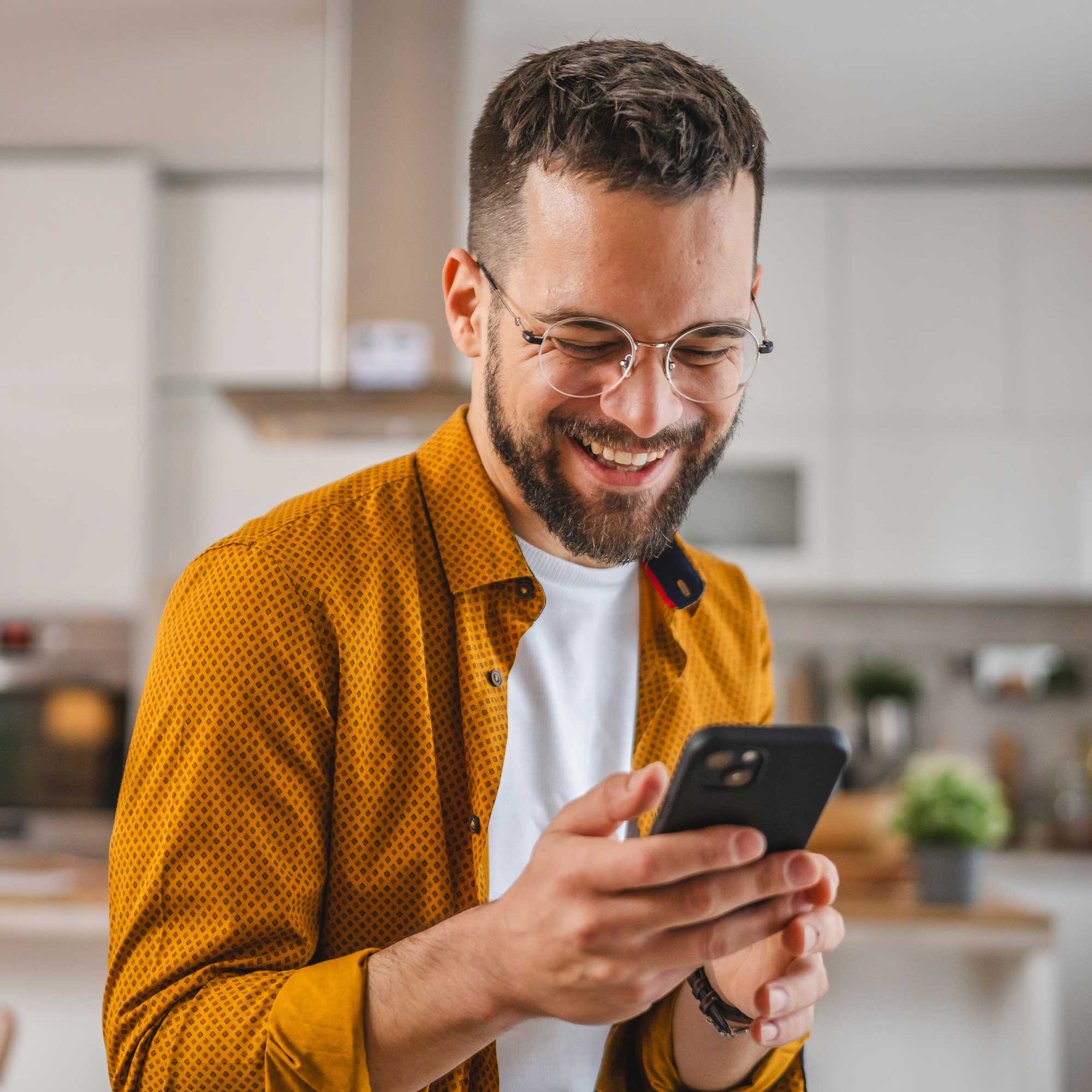 A man wearing a denim shirt smiles as he uses his smartphone.