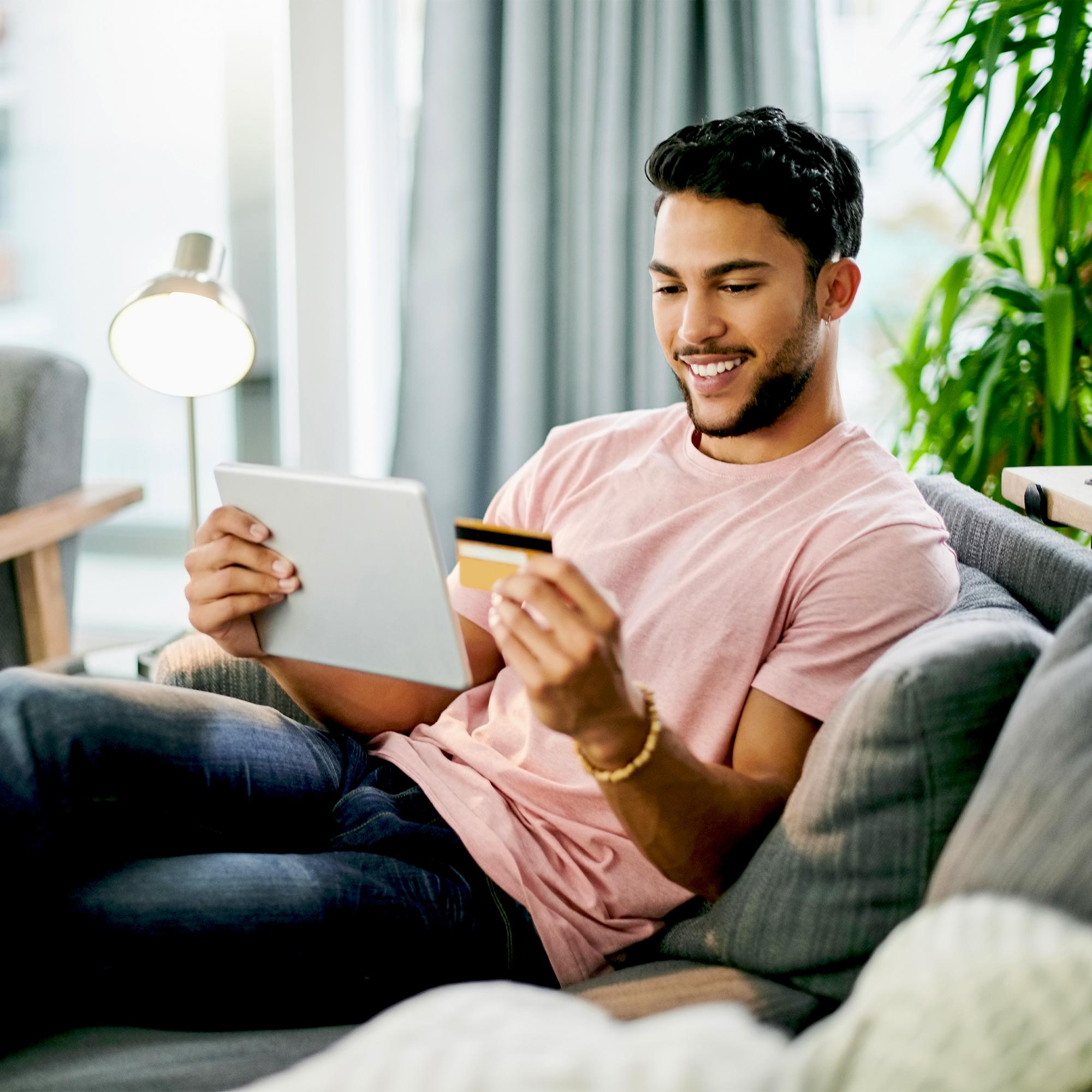 A man sitting on his couch looking at his credit card with ipad in hand.