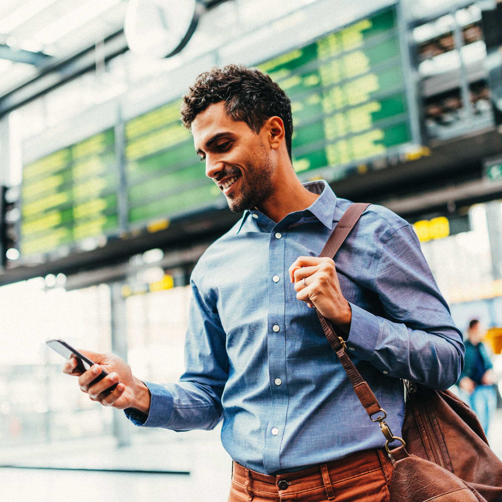 A man carrying a messenger bag, stands in the airport smiling and looking at his phone.