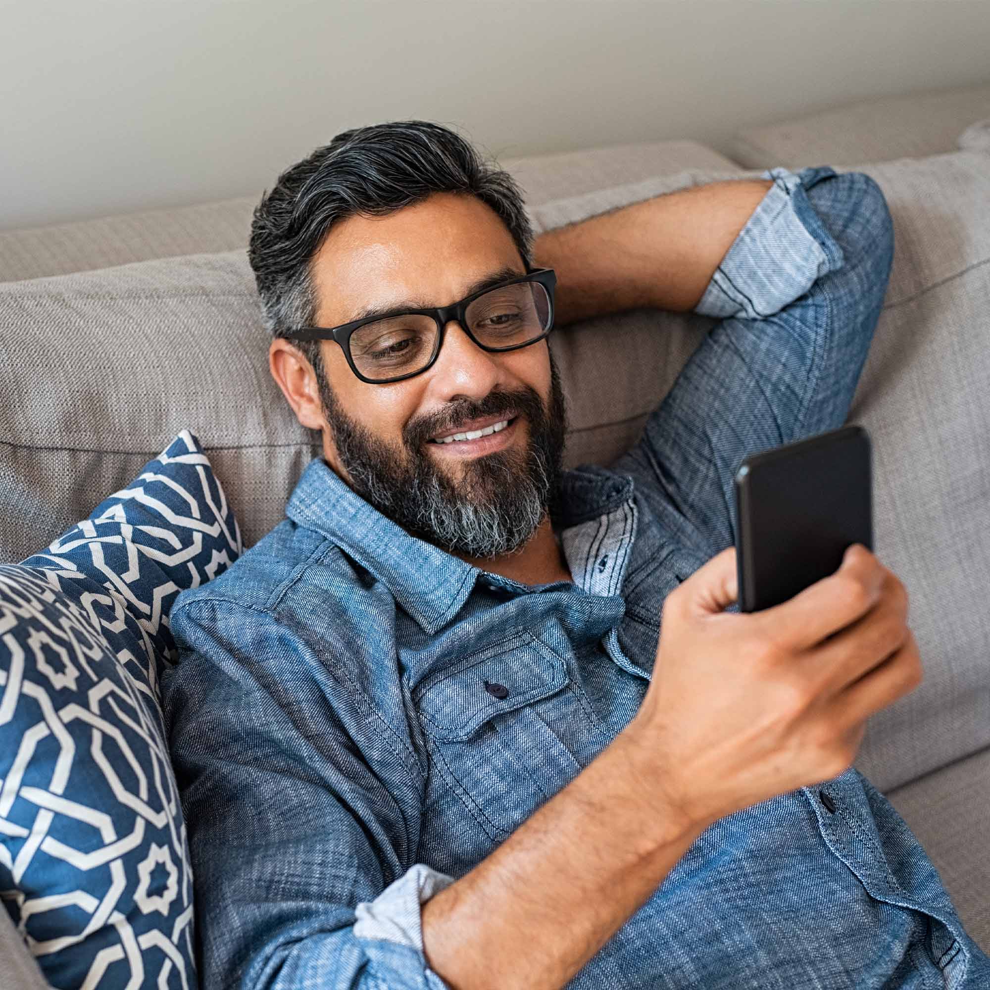 A man wearing a denim shirt smiles as he uses his smartphone.